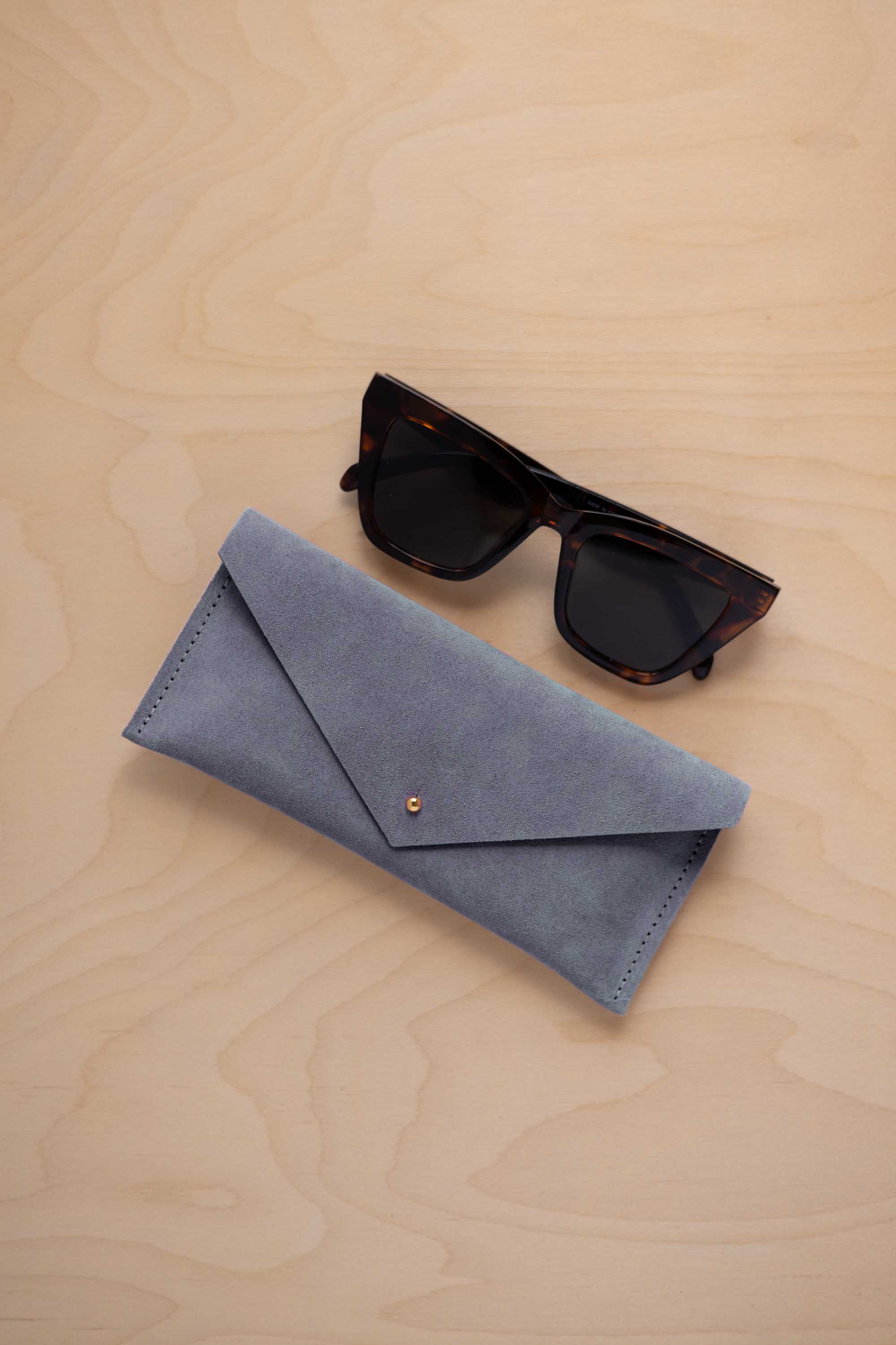 A pair of dark tortoiseshell sunglasses sit next to a dusky blue suede envelope style eye glasses case with a small gold button, set on a light wood grain background.