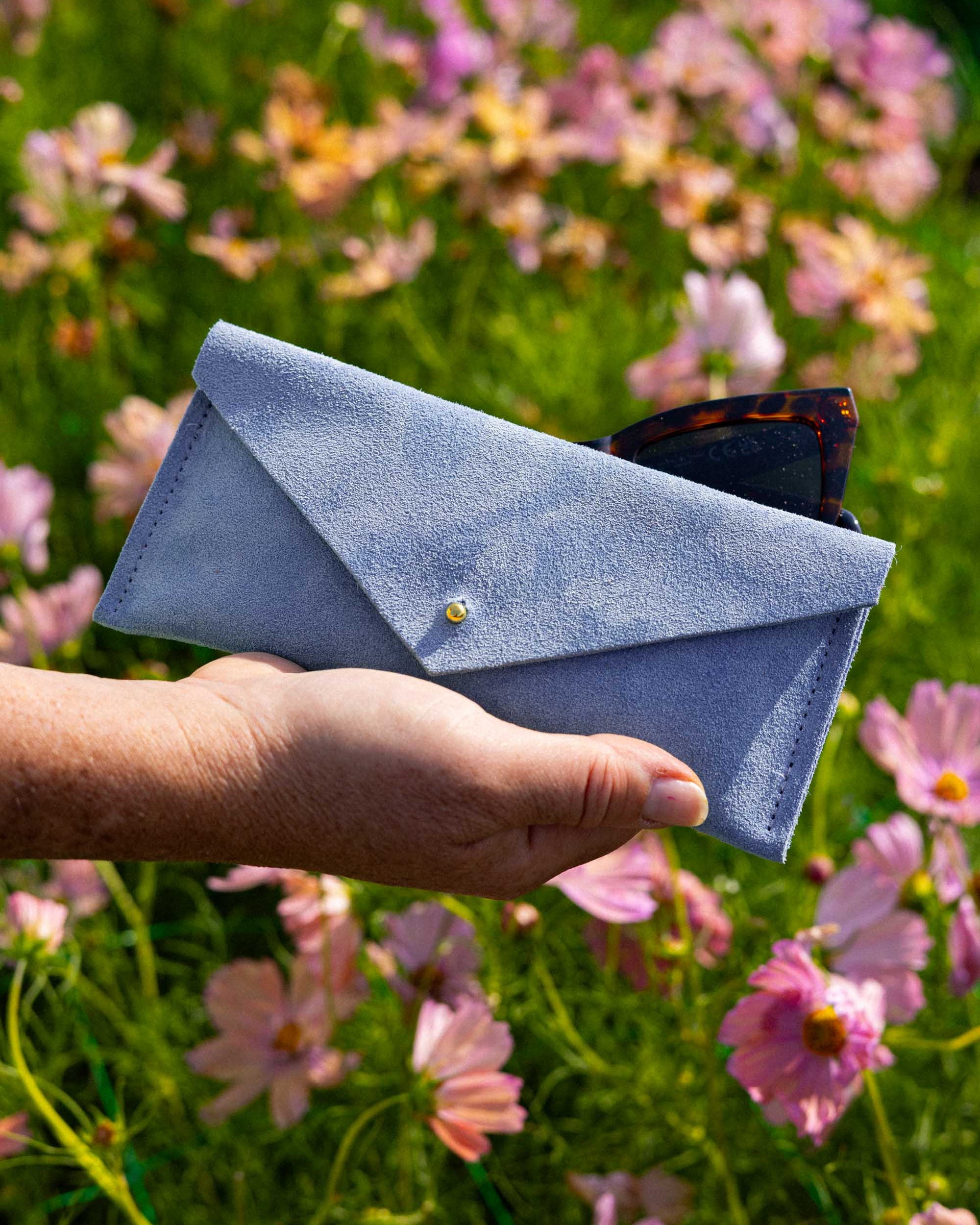 A light blue suede envelope-style eye glasses case with a small gold button, held in a person's hand against a blurred background of pink and purple flowers in a green field.