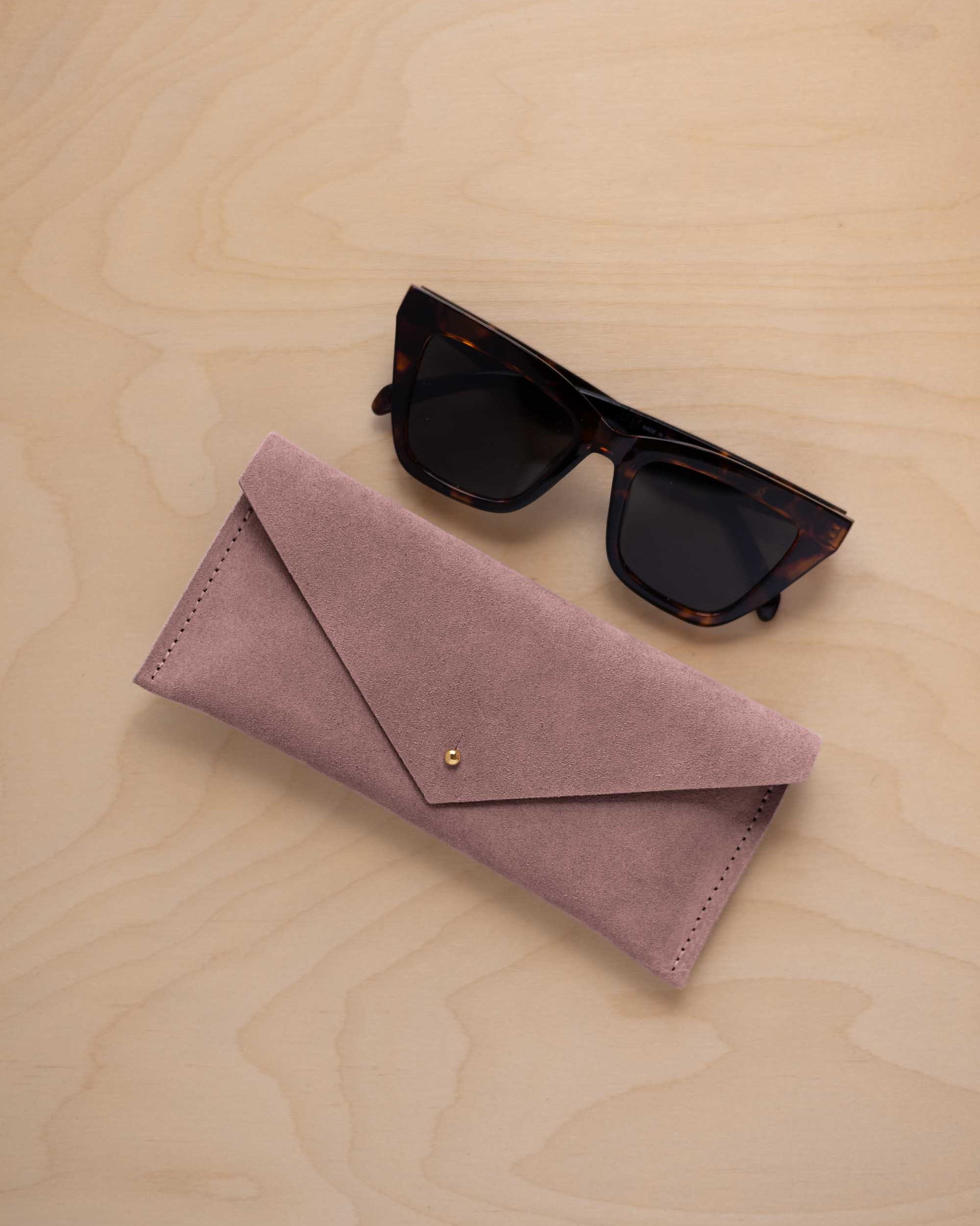 A pair of dark tortoiseshell sunglasses sit next to a dusty mauve grey suede envelope style eye glasses case with a small gold button, set on a light wood grain background.