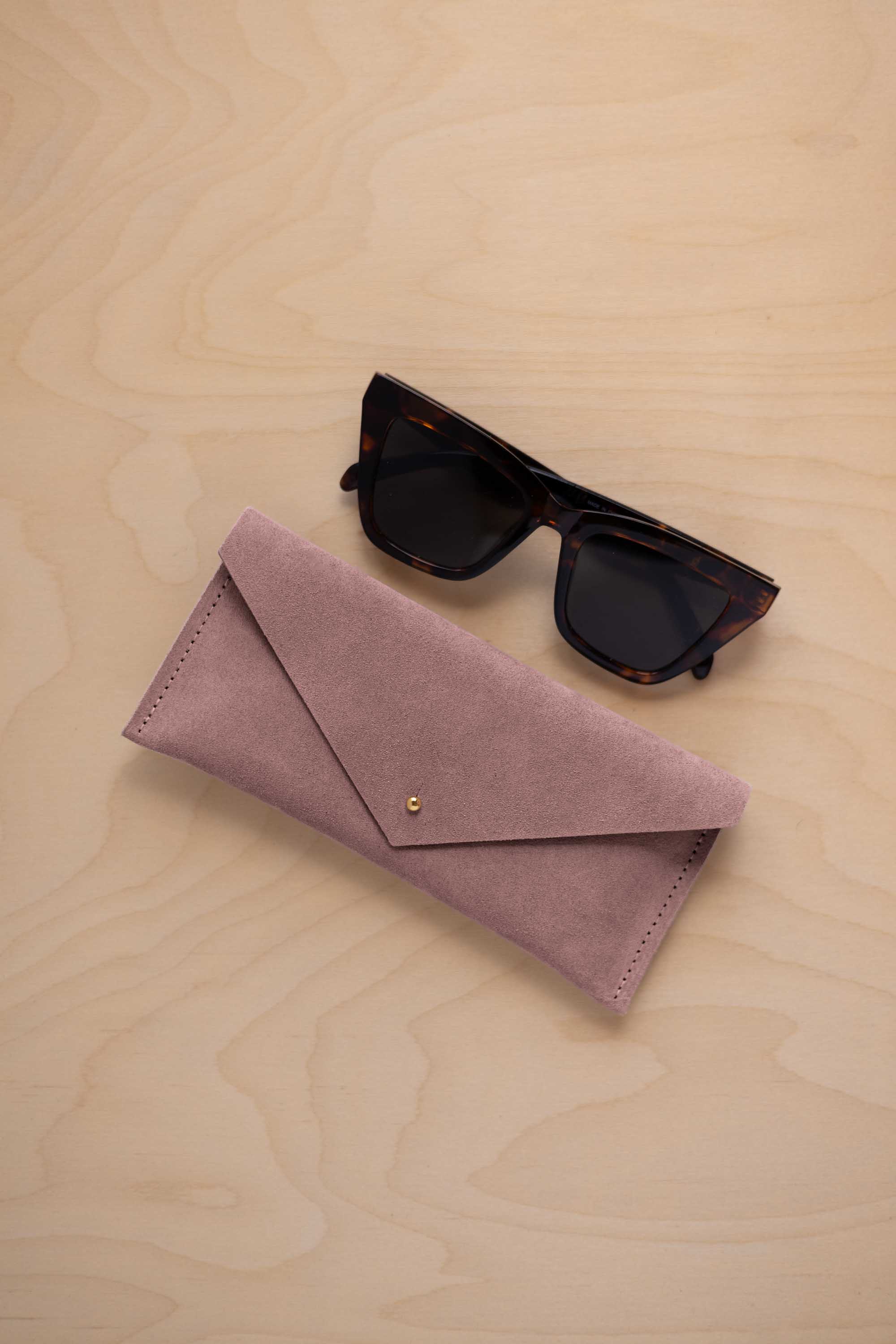 A pair of dark tortoiseshell sunglasses sit next to a dusty mauve grey suede envelope style eye glasses case with a small gold button, set on a light wood grain background.