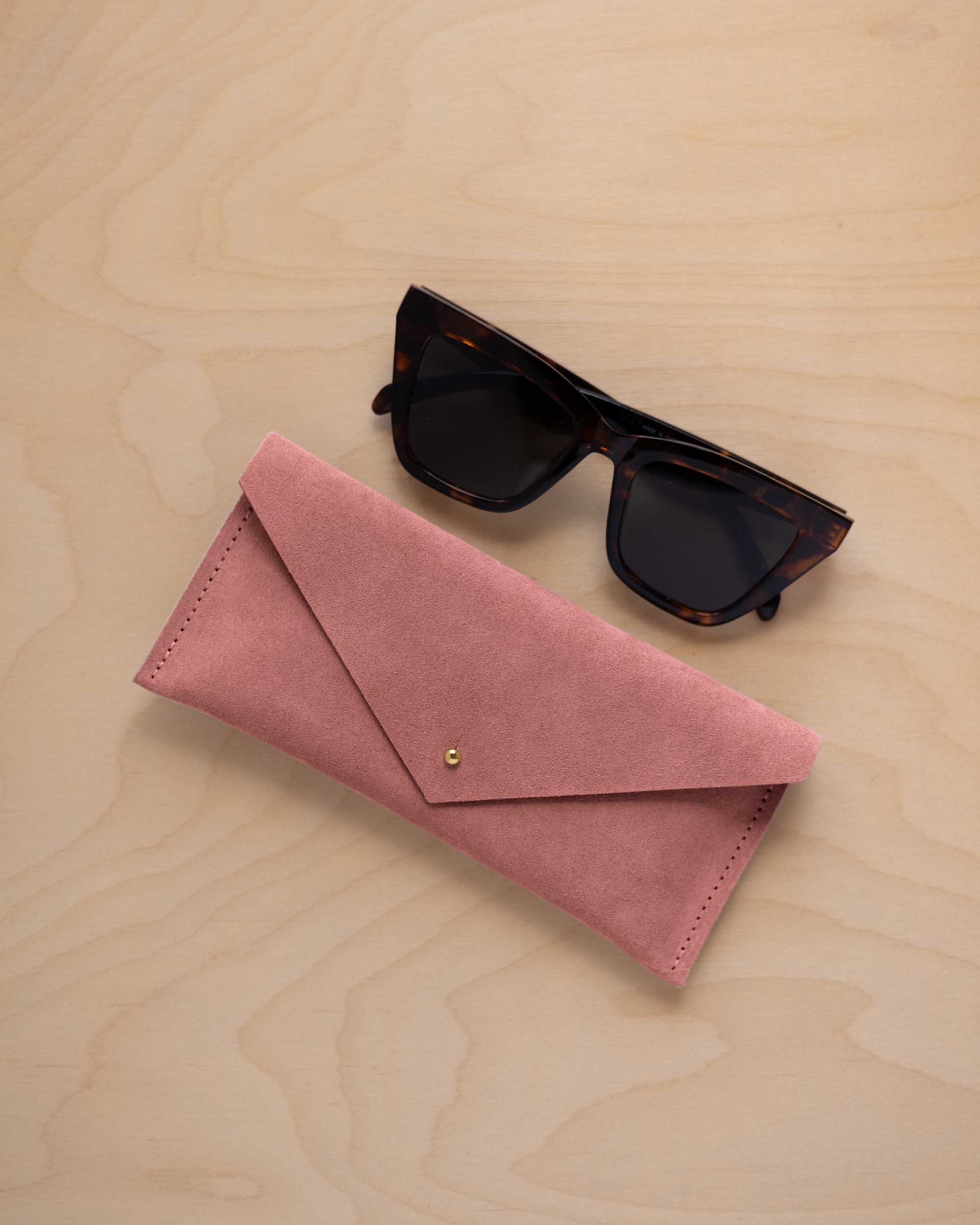 A pair of dark tortoiseshell sunglasses sit next to a dusty rose pink suede envelope style eye glasses case with a small gold button, set on a light wood grain background.