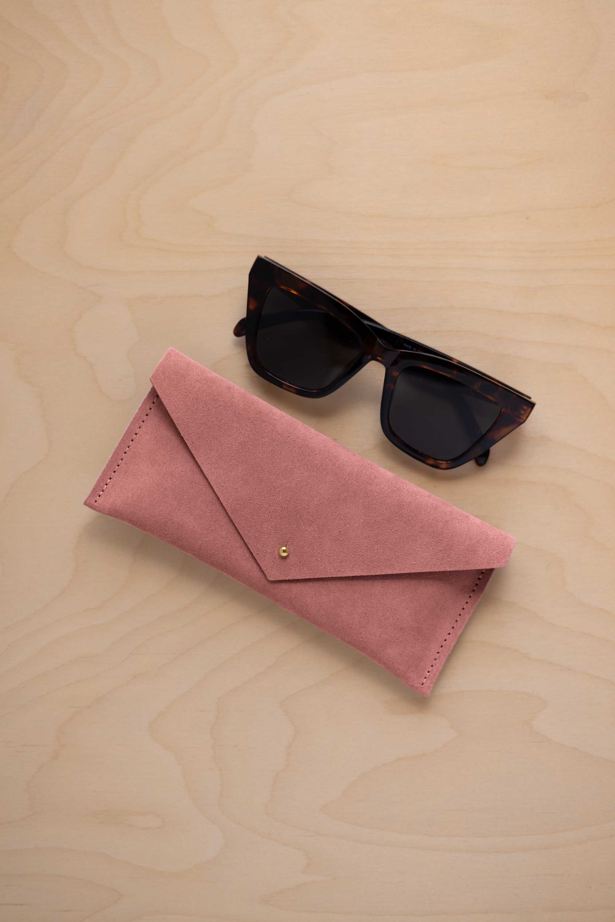 A pair of dark tortoiseshell sunglasses sit next to a dusty rose pink suede envelope style eye glasses case with a small gold button, set on a light wood grain background.