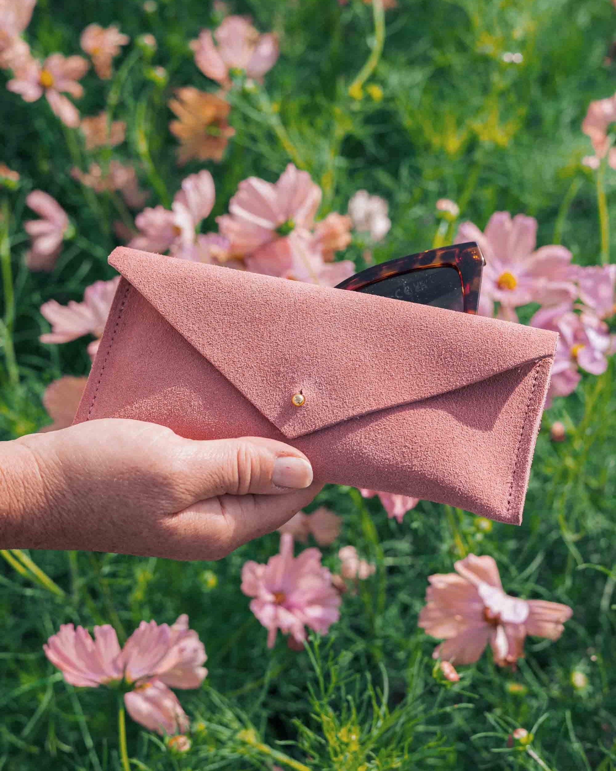 A hand holding a dusty pink envelope-style suede eyeglasses case with a small gold button, set against a blurred background of pink flowers and green foliage.