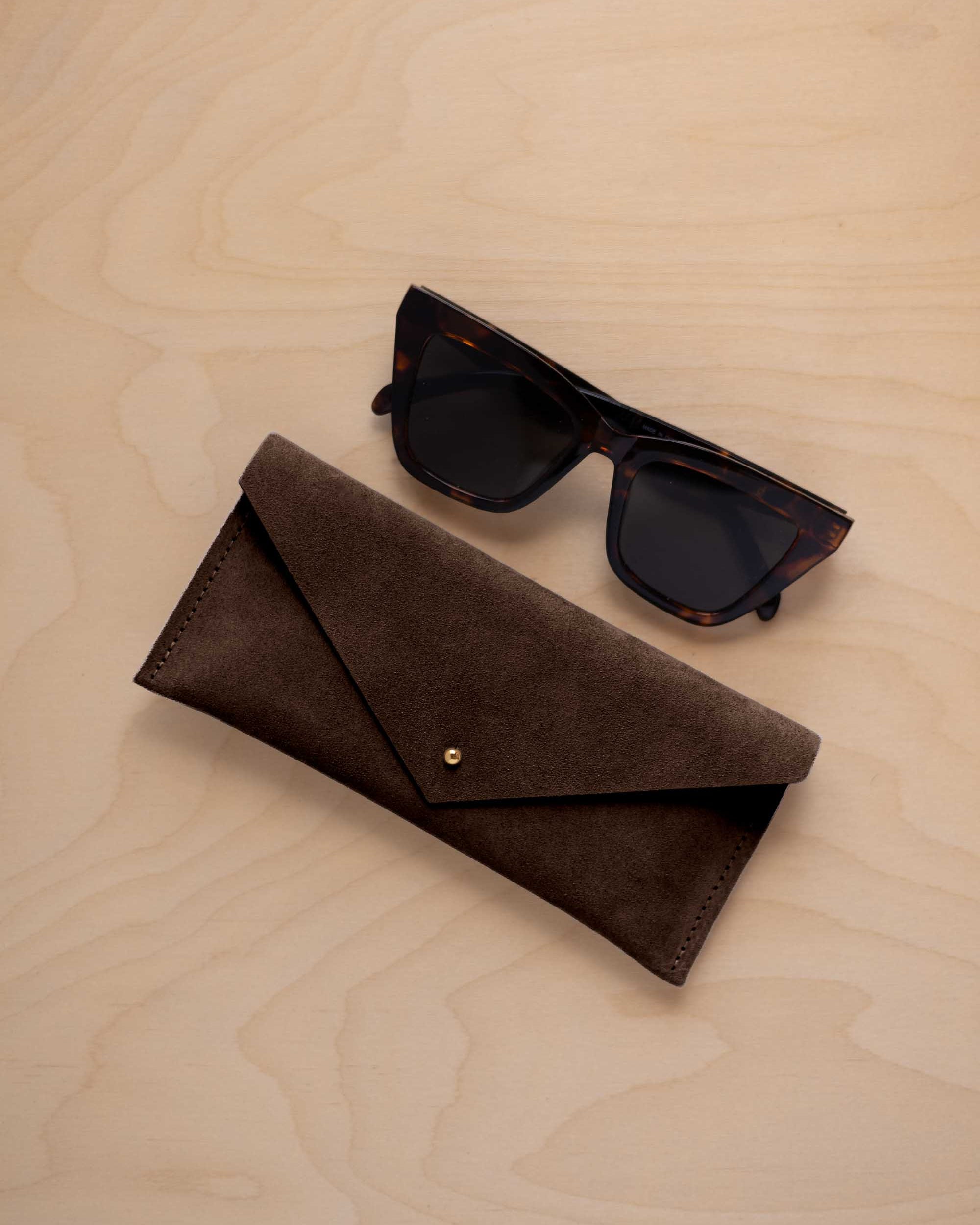 A pair of dark tortoiseshell sunglasses sit next to an espresso brown coloured suede envelope style eye glasses case with a small gold button, set on a light wood grain background.