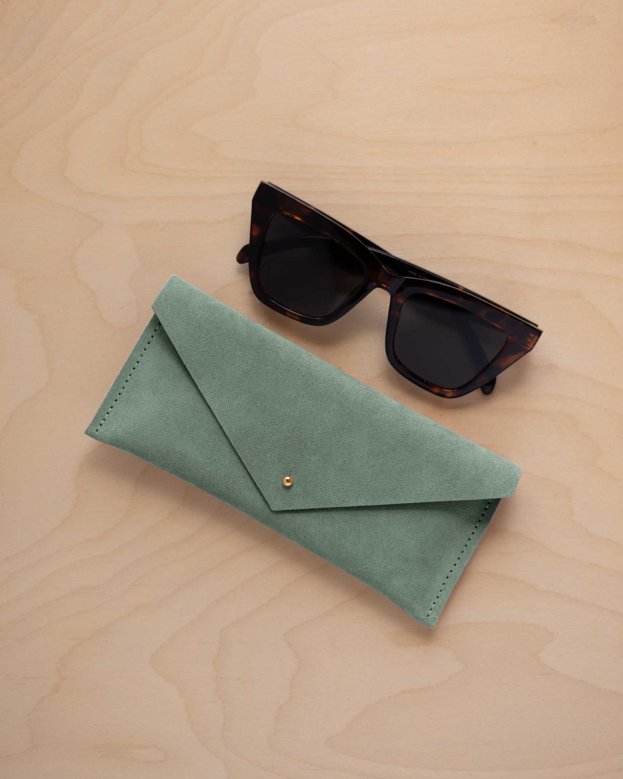 A pair of dark tortoiseshell sunglasses sit next to a eucalyptus sage green coloured suede envelope style eye glasses case with a small gold button, set on a light wood grain background.