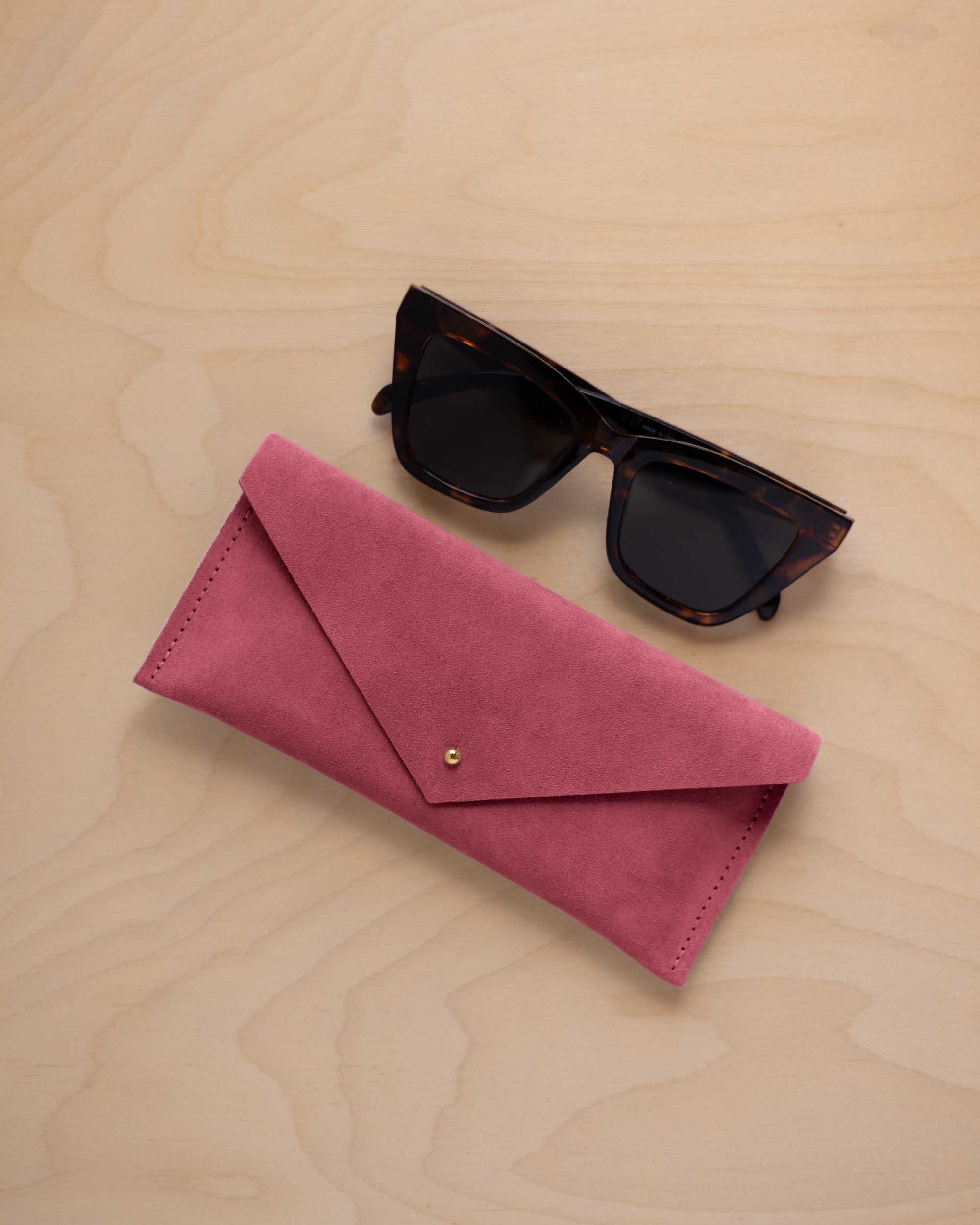 A pair of dark tortoiseshell sunglasses sit next to a faded rose pink suede envelope style eye glasses case with a small gold button, set on a light wood grain background.
