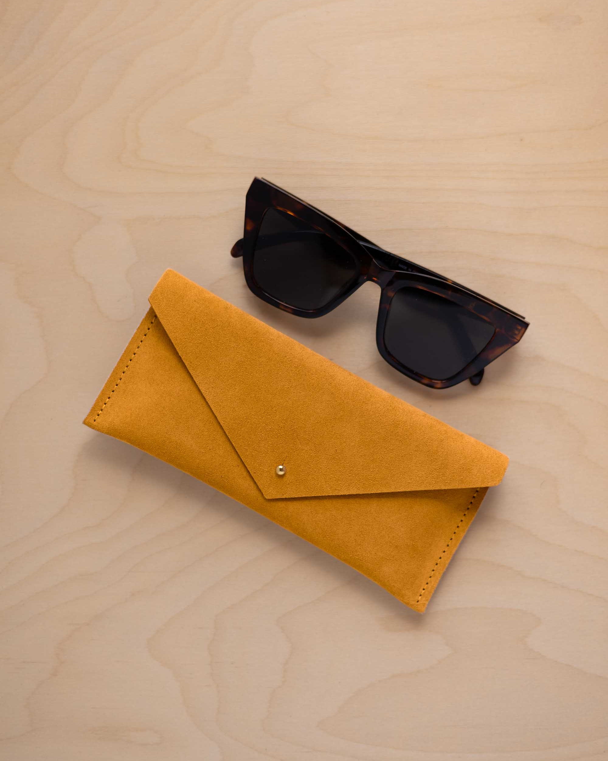 A pair of dark tortoiseshell sunglasses sit next to a mustard yellow suede envelope style eye glasses case with a small gold button, set on a light wood grain background.