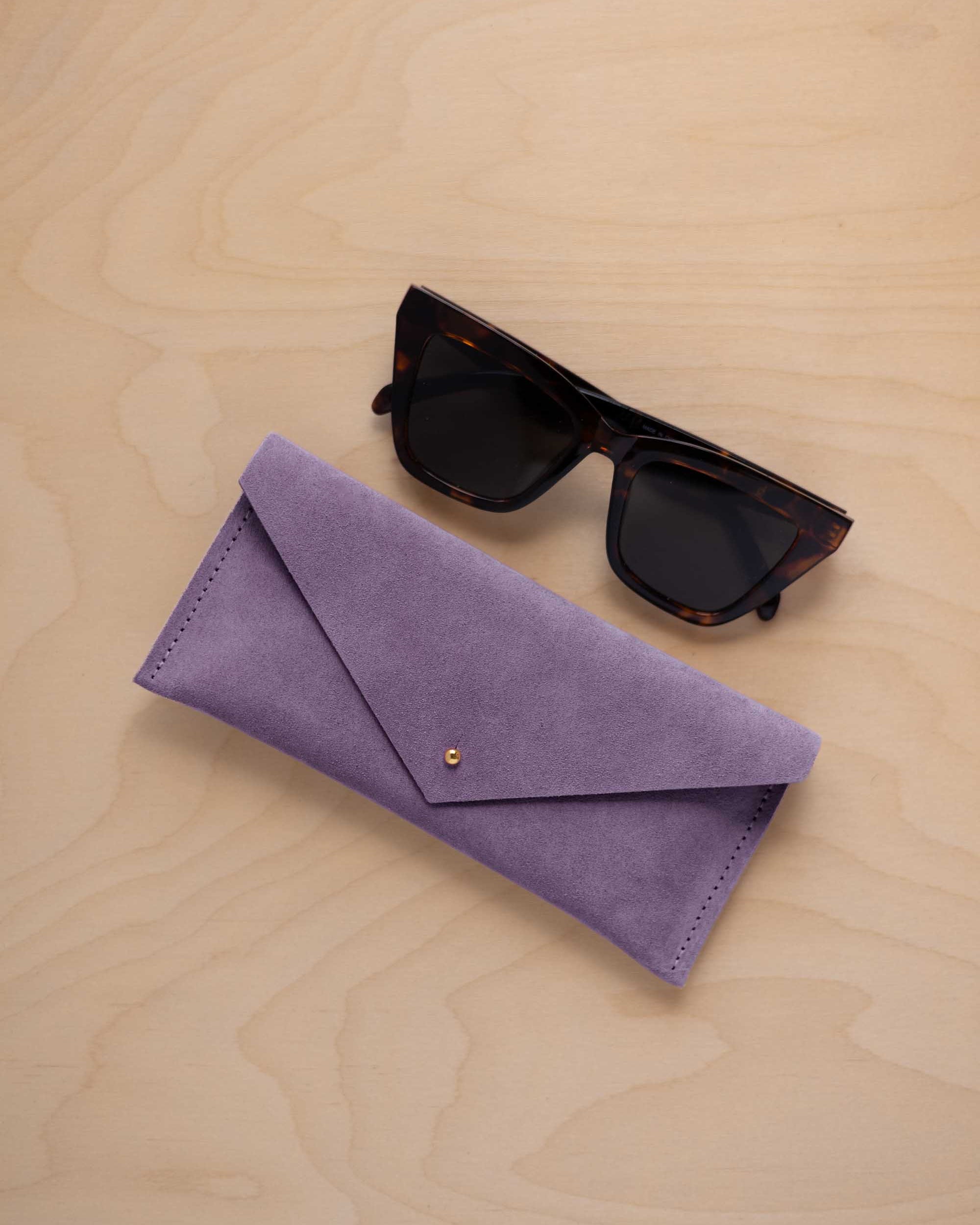 A pair of dark tortoiseshell sunglasses sit next to a lavender suede envelope style eye glasses case with a small gold button, set on a light wood grain background.