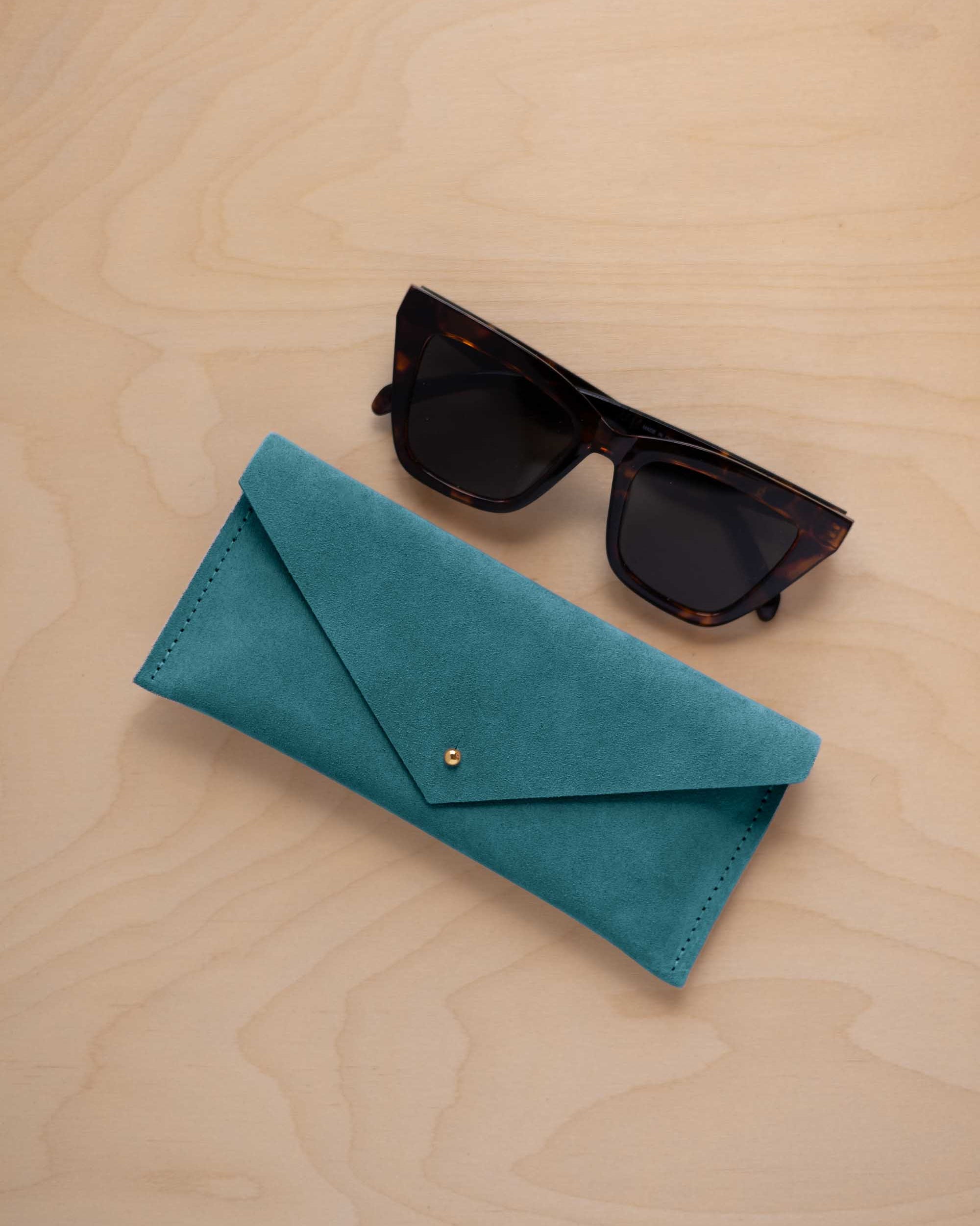 A pair of dark tortoiseshell sunglasses sit next to a marine teal coloured suede envelope style eye glasses case with a small gold button, set on a light wood grain background.