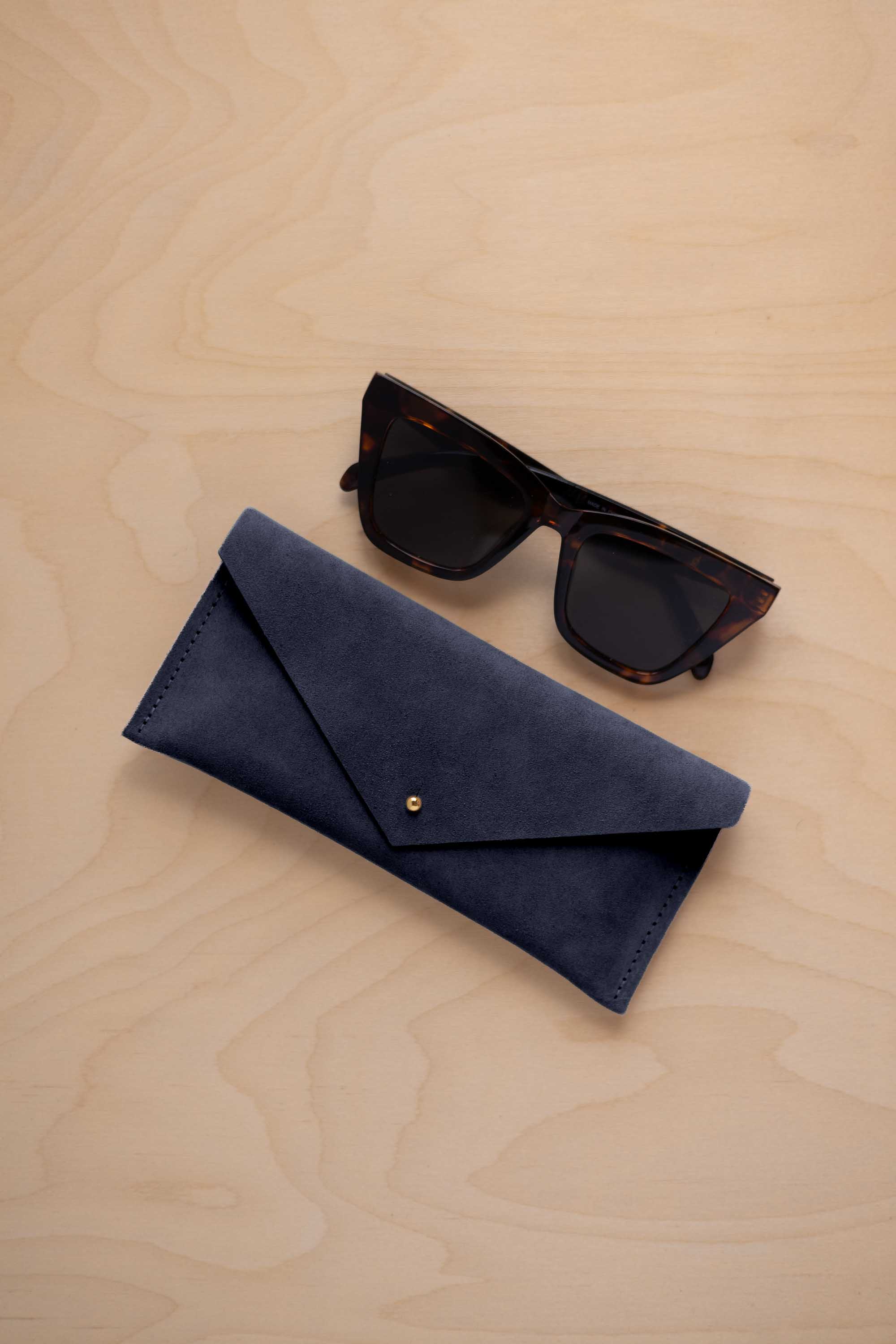 A pair of dark tortoiseshell sunglasses sit next to a midnight navy coloured suede envelope style eye glasses case with a small gold button, set on a light wood grain background.