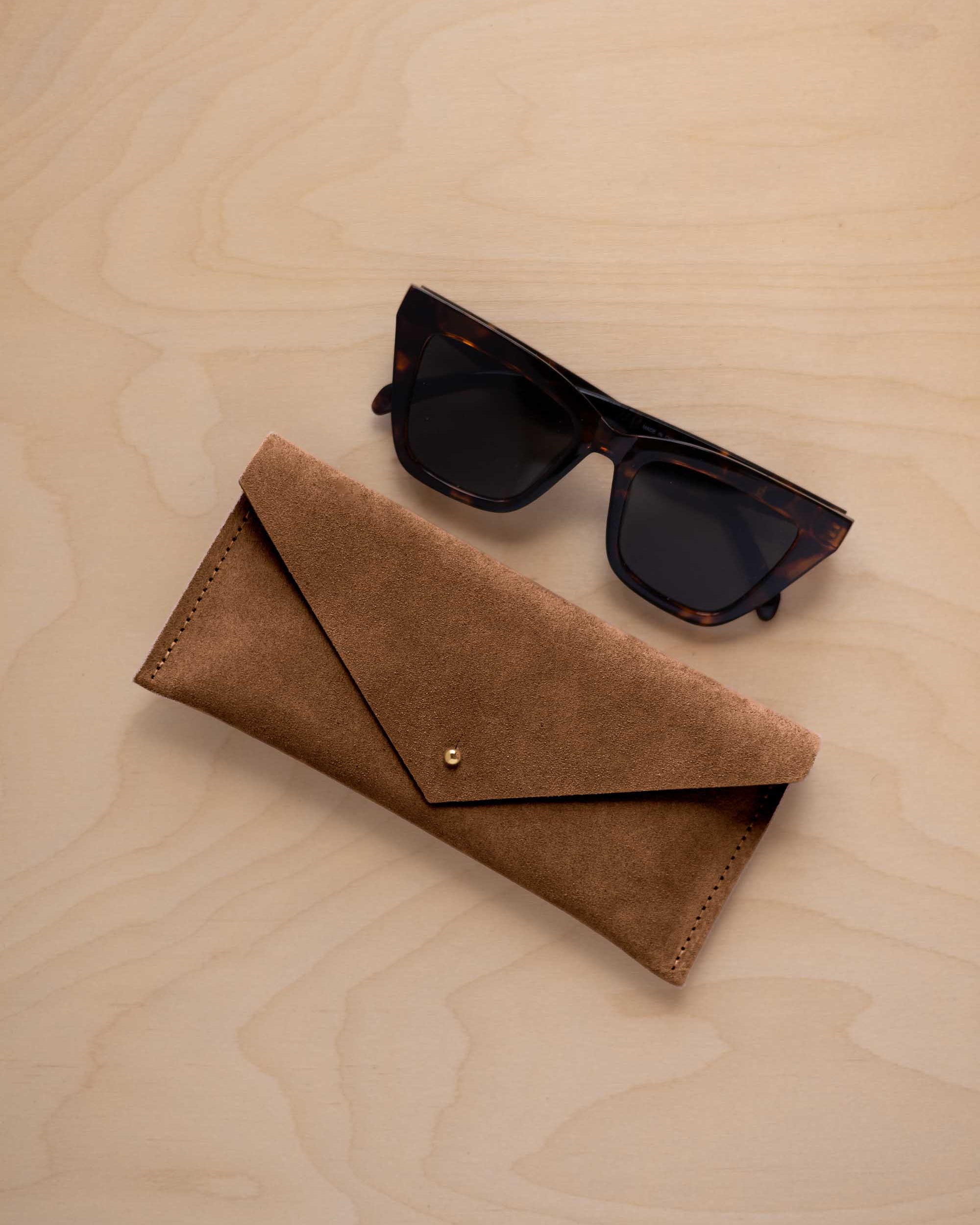 A pair of dark tortoiseshell sunglasses sit next to a mocha brown suede envelope style eye glasses case with a small gold button, set on a light wood grain background.