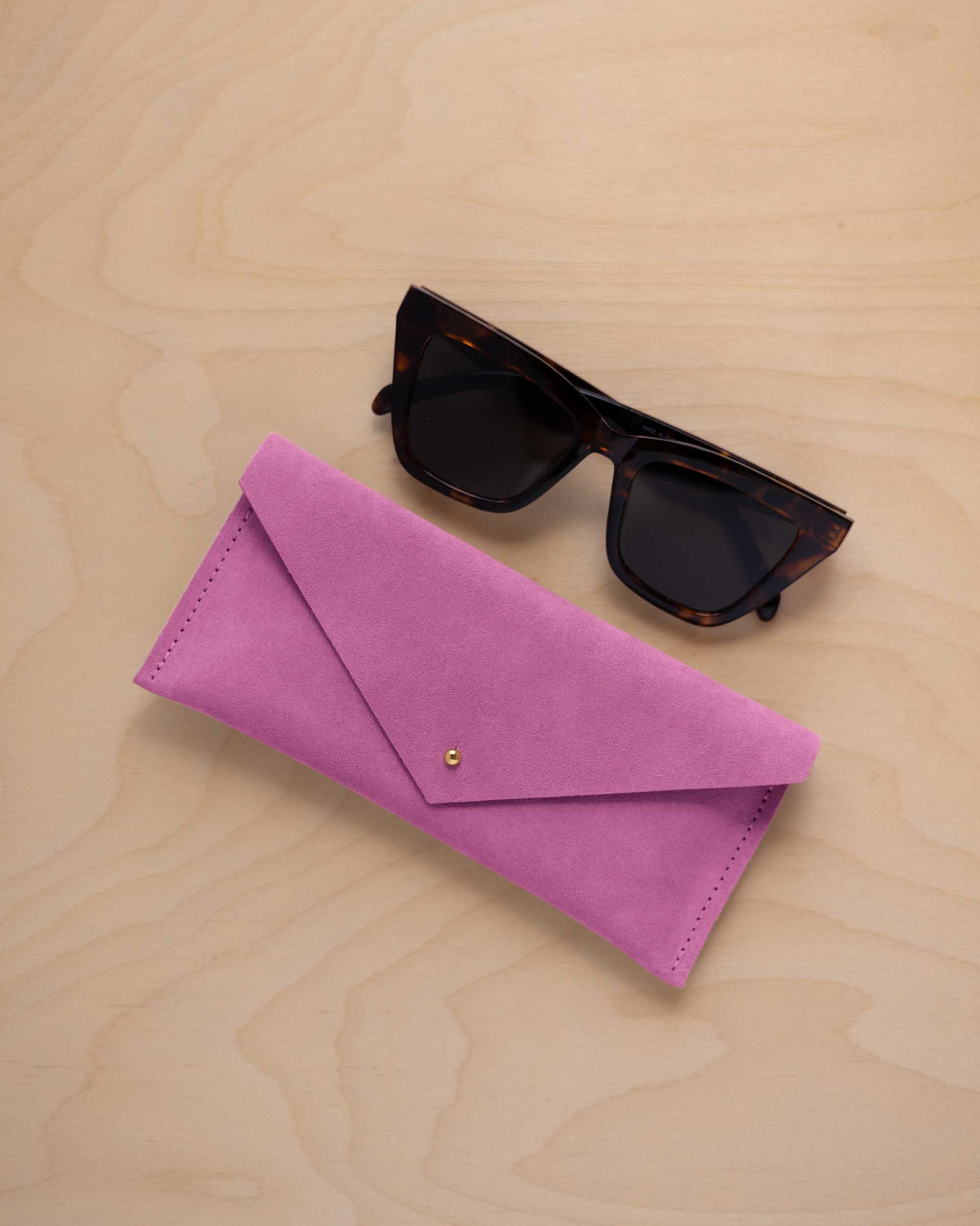 A pair of dark tortoiseshell sunglasses sit next to a peony pink suede envelope style eye glasses case with a small gold button, set on a light wood grain background.