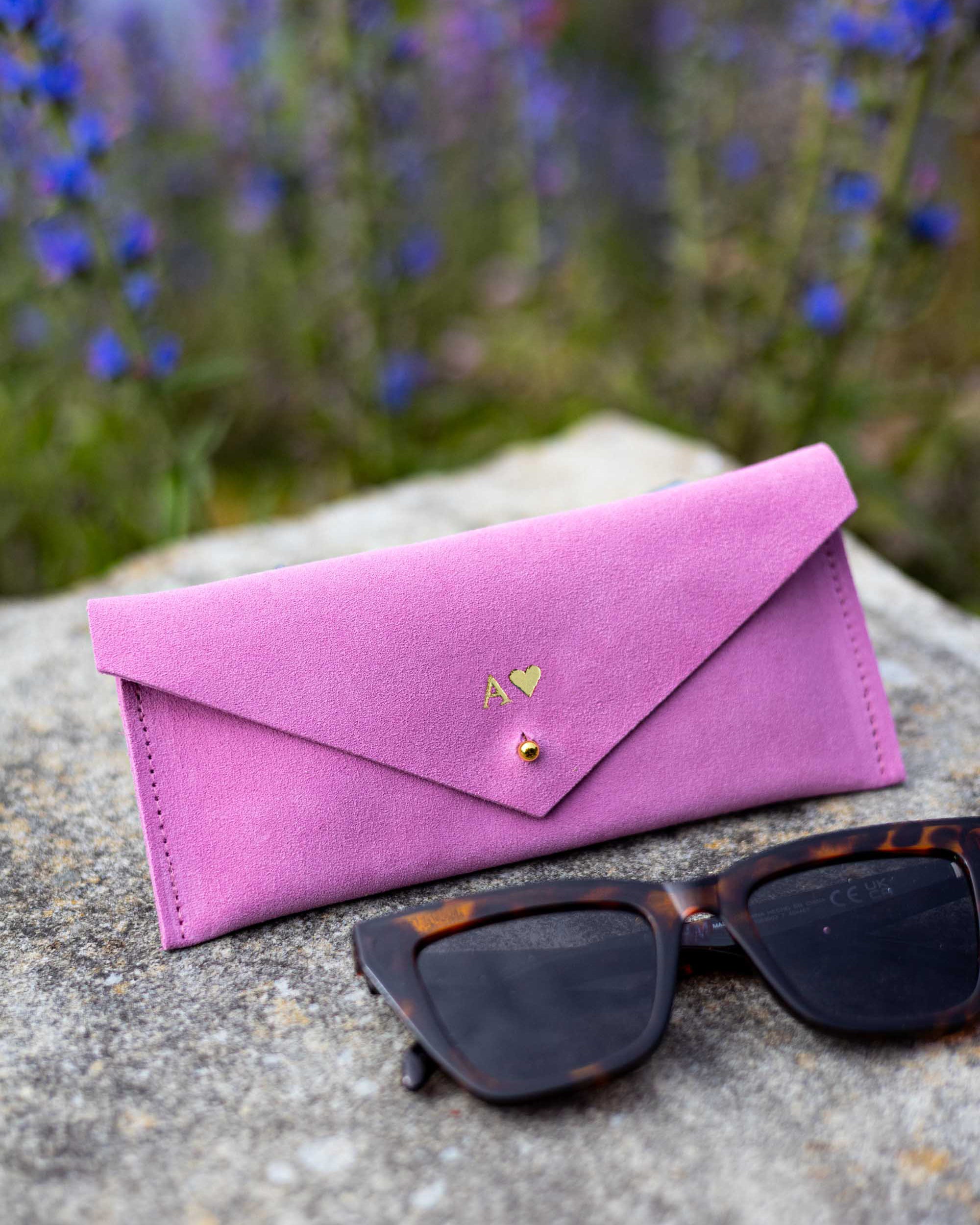 A pink suede envelope-style sunglasses case personalised with a small gold heart logo and the initial A, placed on a stone surface outdoors with tortoiseshell sunglasses in front and blurred blue flowers in the background.