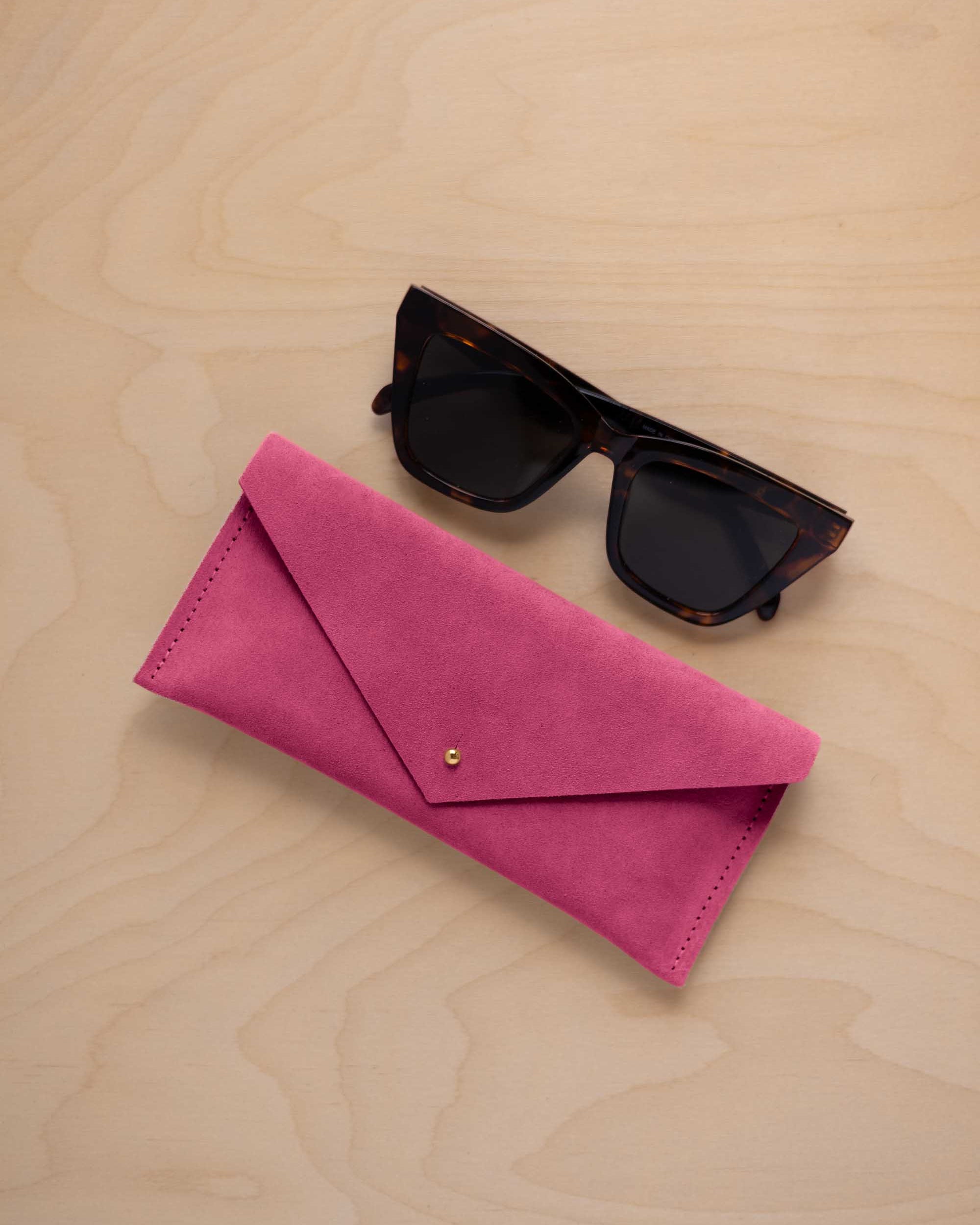 A pair of dark tortoiseshell sunglasses sit next to a raspberry pink suede envelope style eye glasses case with a small gold button, set on a light wood grain background.