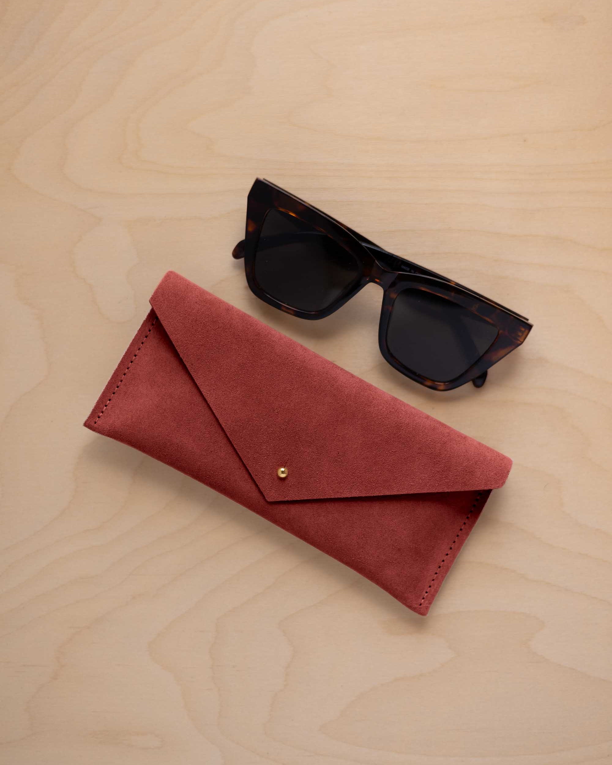 A pair of dark tortoiseshell sunglasses sit next to a rust red suede envelope style eye glasses case with a small gold button, set on a light wood grain background.