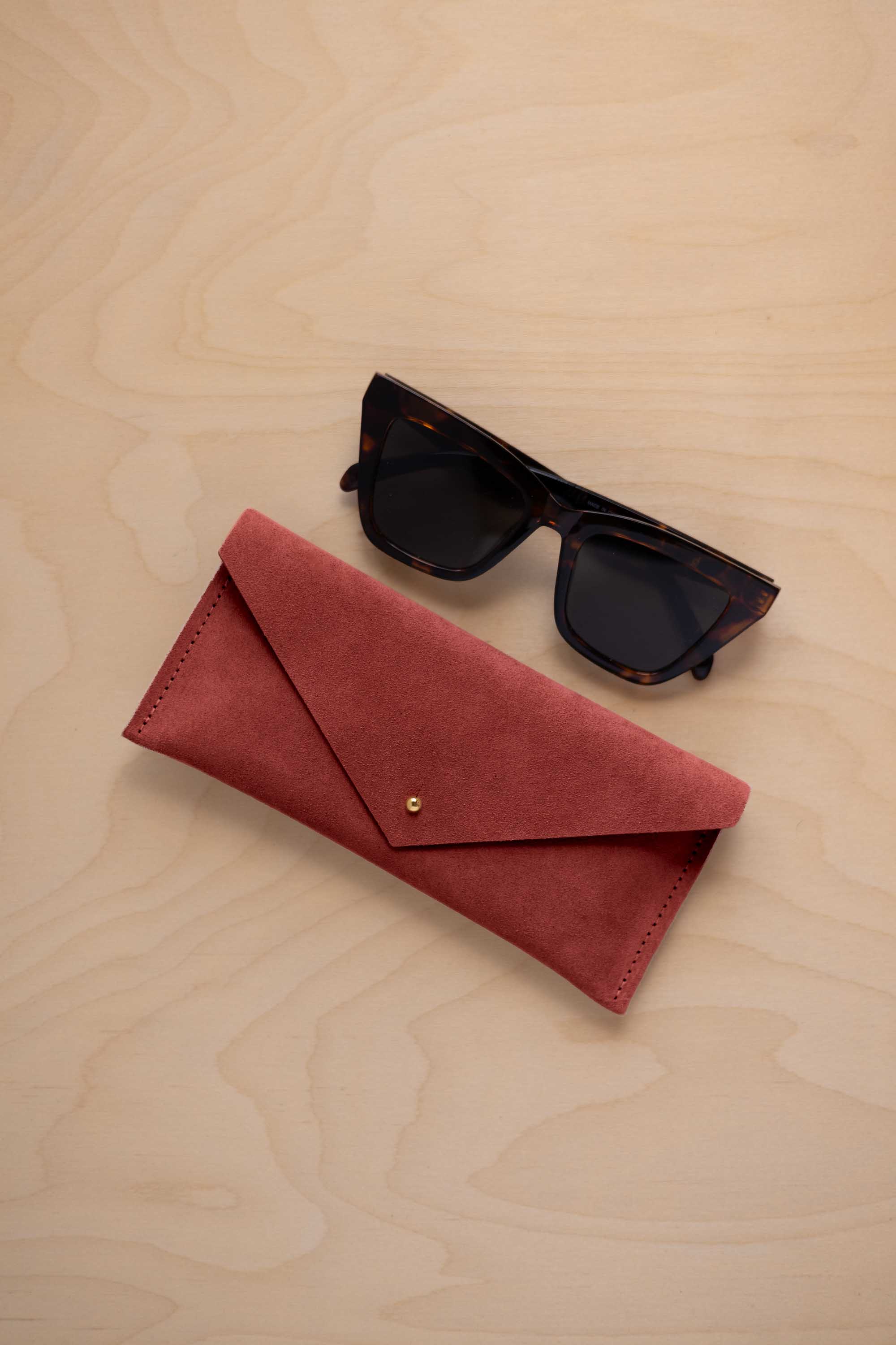 A pair of dark tortoiseshell sunglasses sit next to a rust red suede envelope style eye glasses case with a small gold button, set on a light wood grain background.
