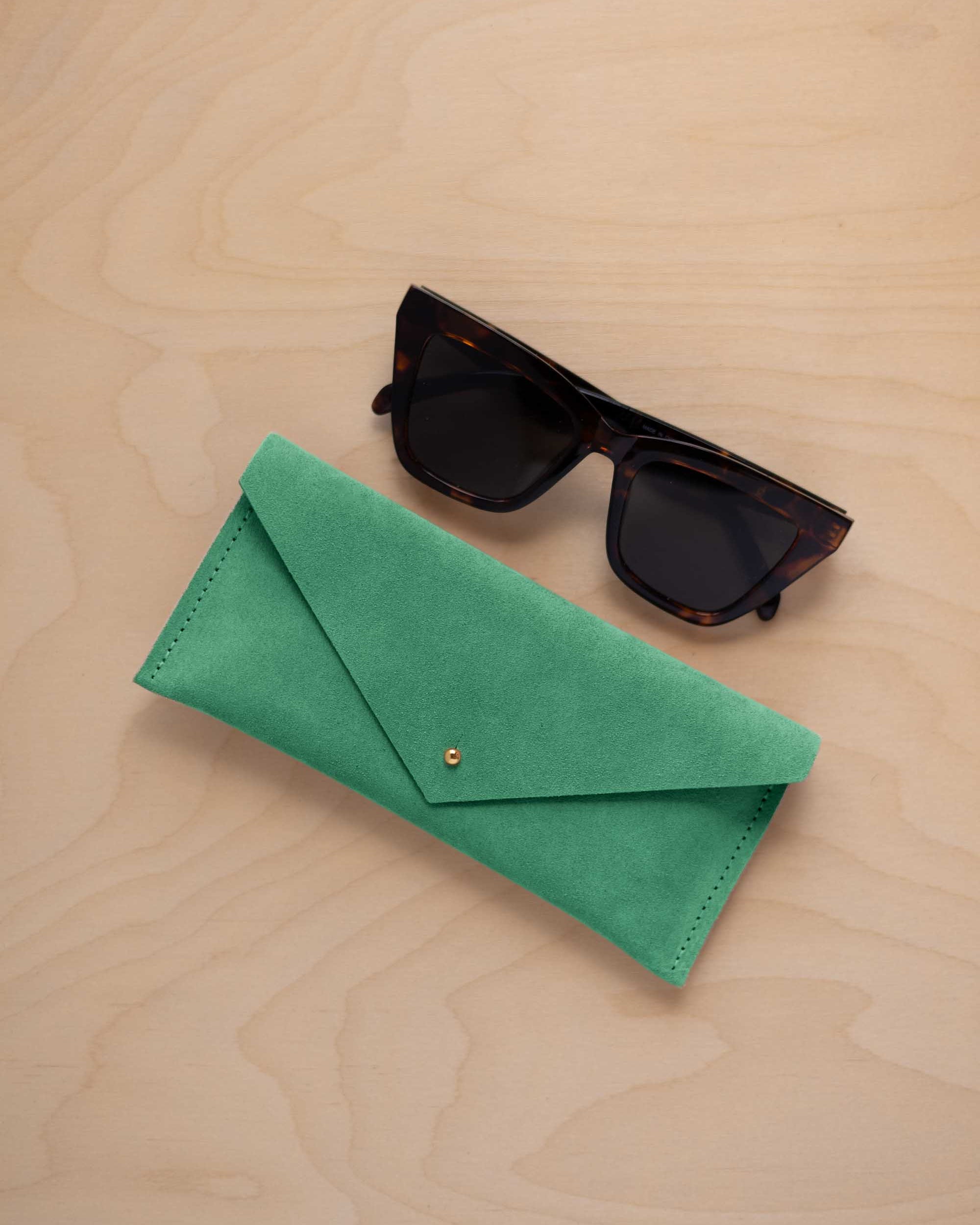 A pair of dark tortoiseshell sunglasses sit next to a seafoam turquoise green coloured suede envelope style eye glasses case with a small gold button, set on a light wood grain background.