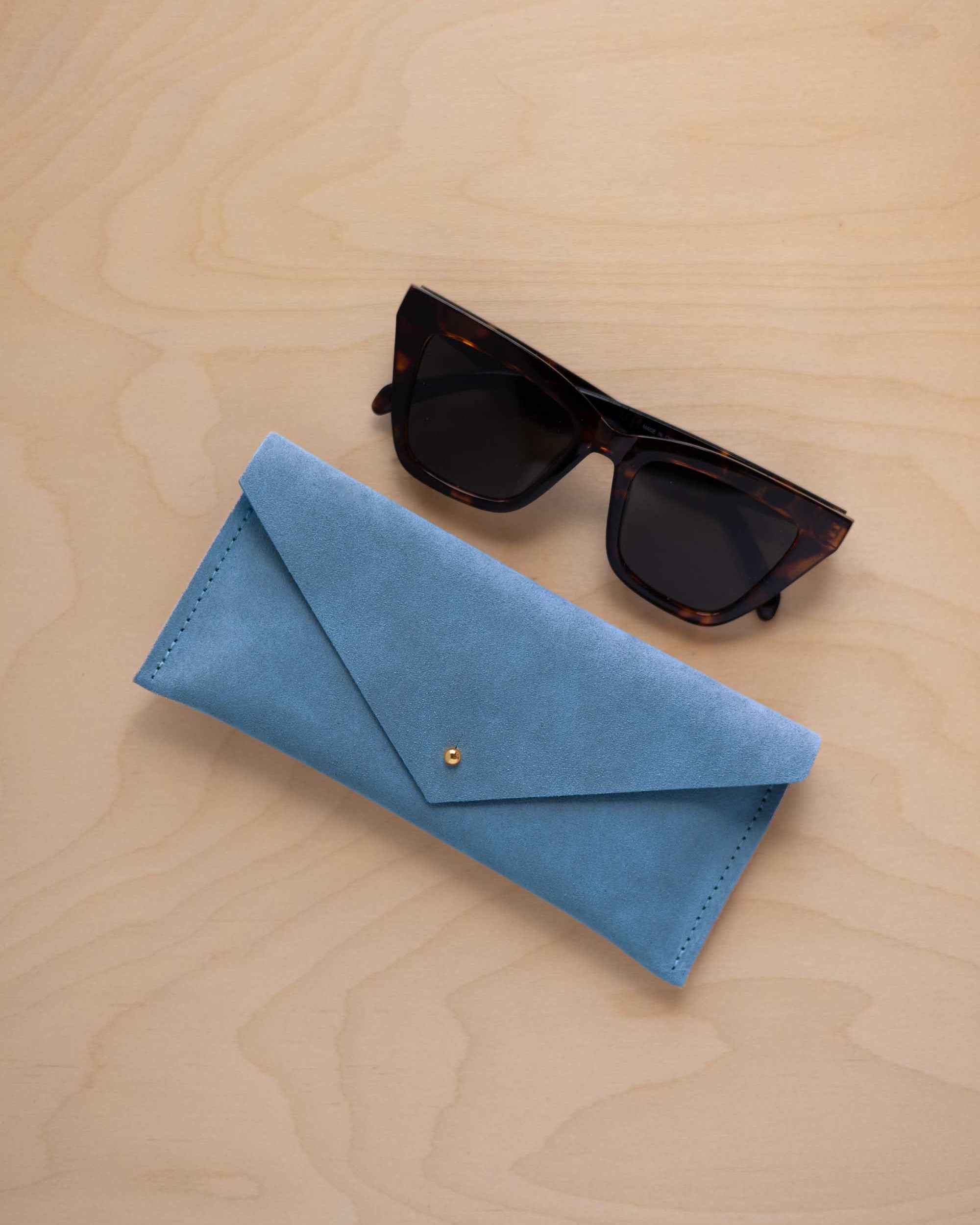 A pair of dark tortoiseshell sunglasses sit next to a sky blue suede envelope style eye glasses case with a small gold button, set on a light wood grain background.