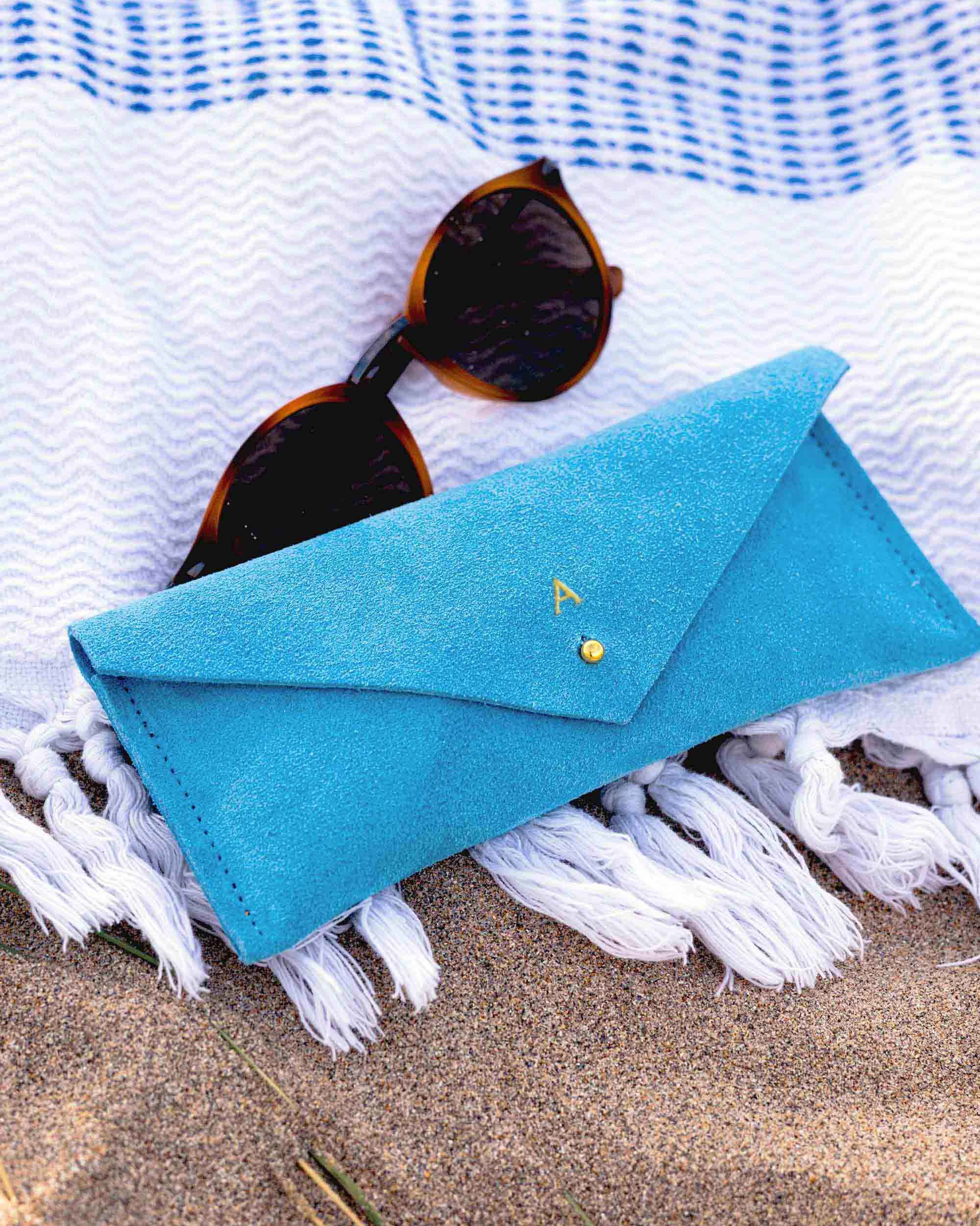 A bright blue envelope-style sunglasses case with a small gold button, styled on a white fringed blanket over sand with tortoiseshell sunglasses resting on top.