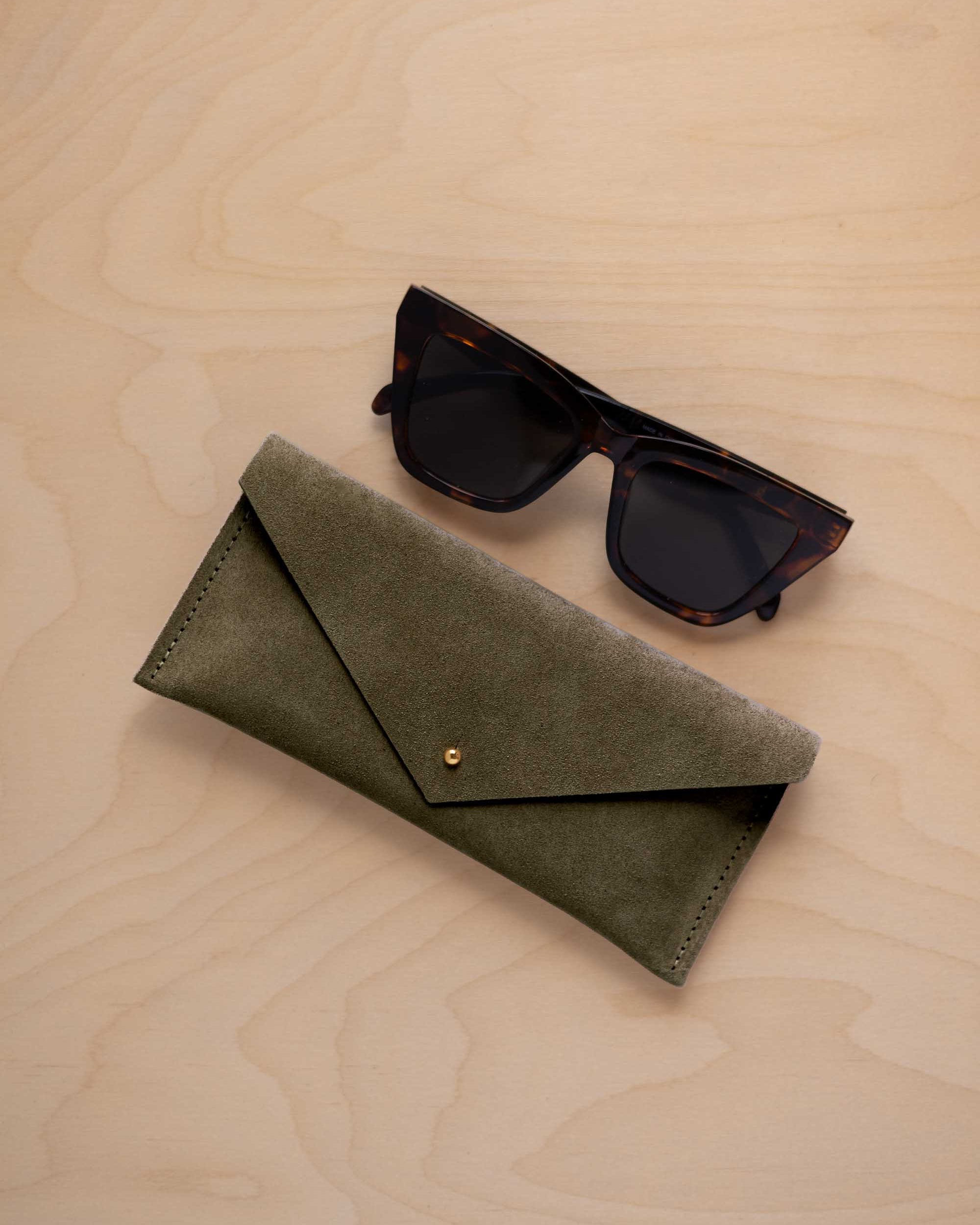 A pair of dark tortoiseshell sunglasses sit next to a soft olive green coloured suede envelope style eye glasses case with a small gold button, set on a light wood grain background.
