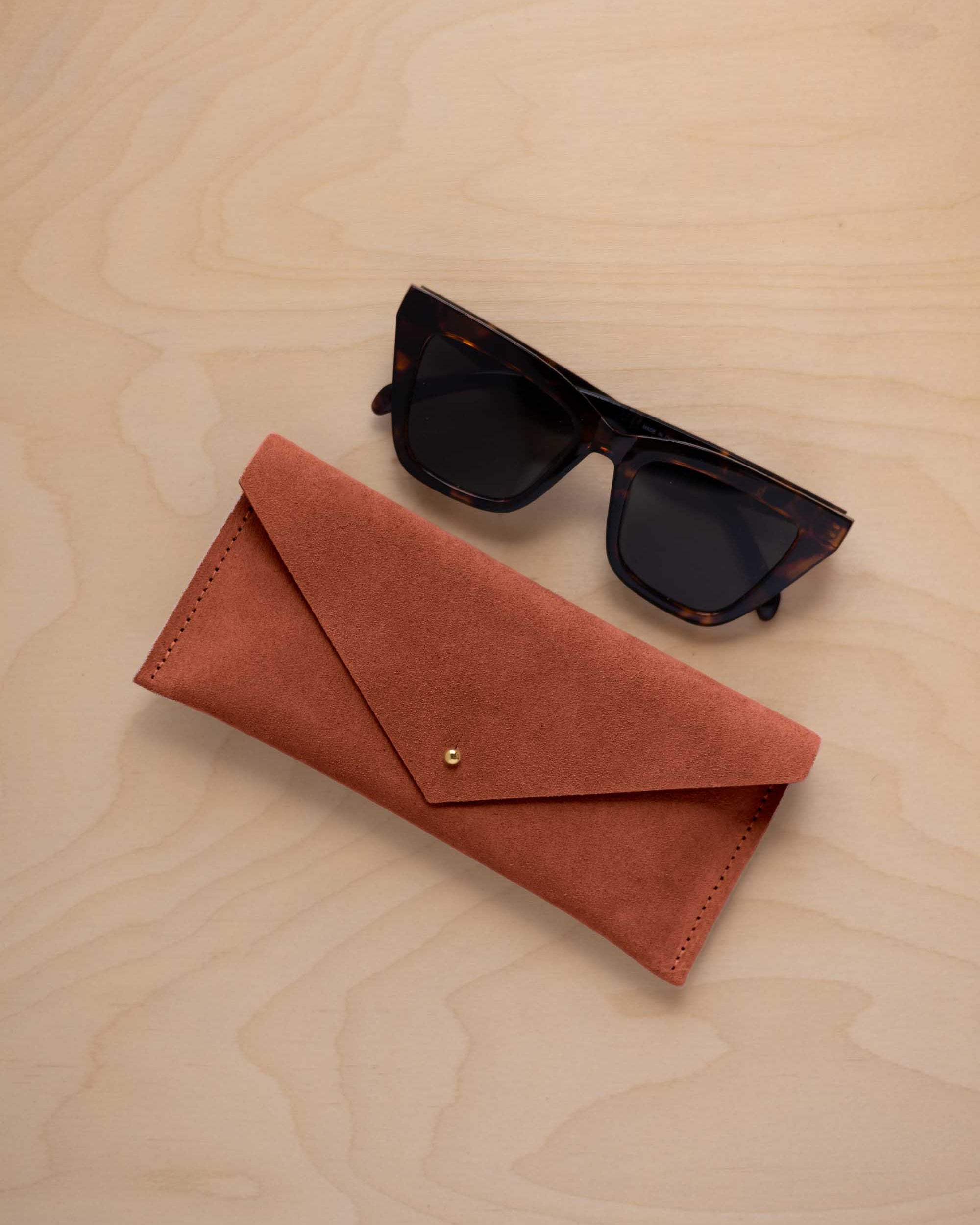 A pair of dark tortoiseshell sunglasses sit next to a terracotta coloured suede envelope style eye glasses case with a small gold button, set on a light wood grain background.
