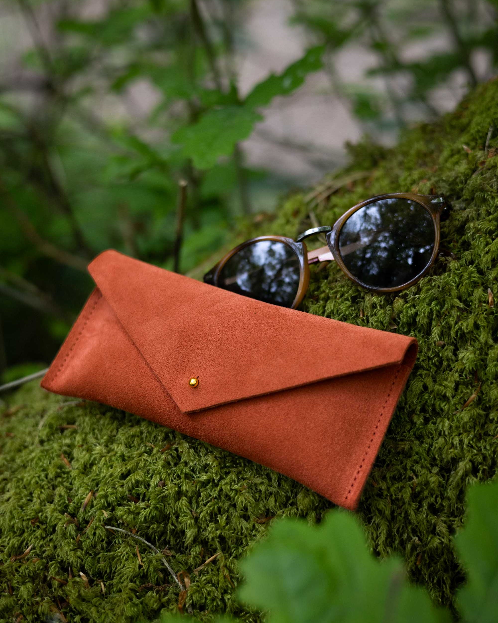 A terracotta orange suede envelope-style sunglasses case with a small gold stud closure, shown outdoors on mossy green foliage with a pair of sunglasses resting on top.