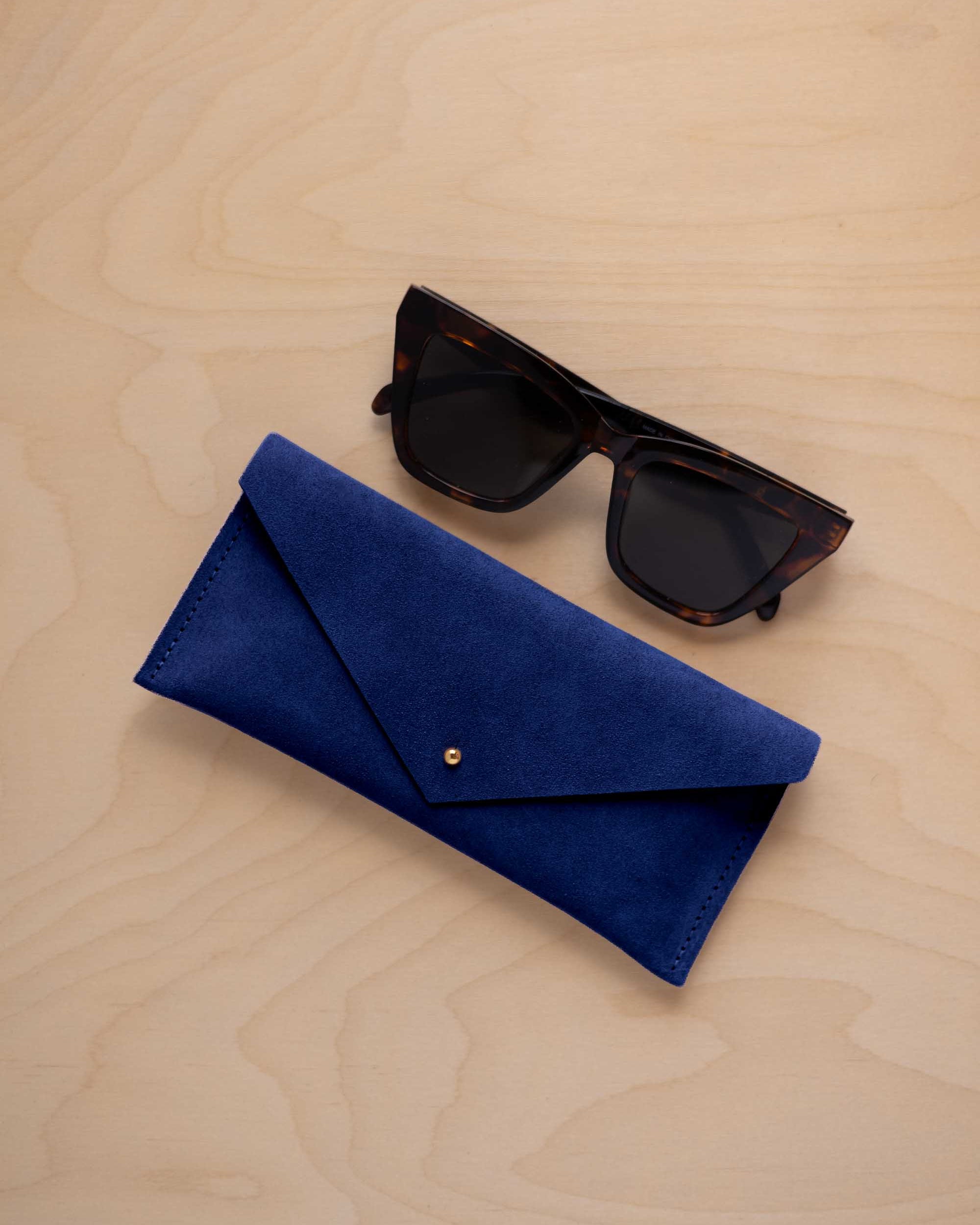 A pair of dark tortoiseshell sunglasses sit next to a ultramarine blue suede envelope style eye glasses case with a small gold button, set on a light wood grain background.