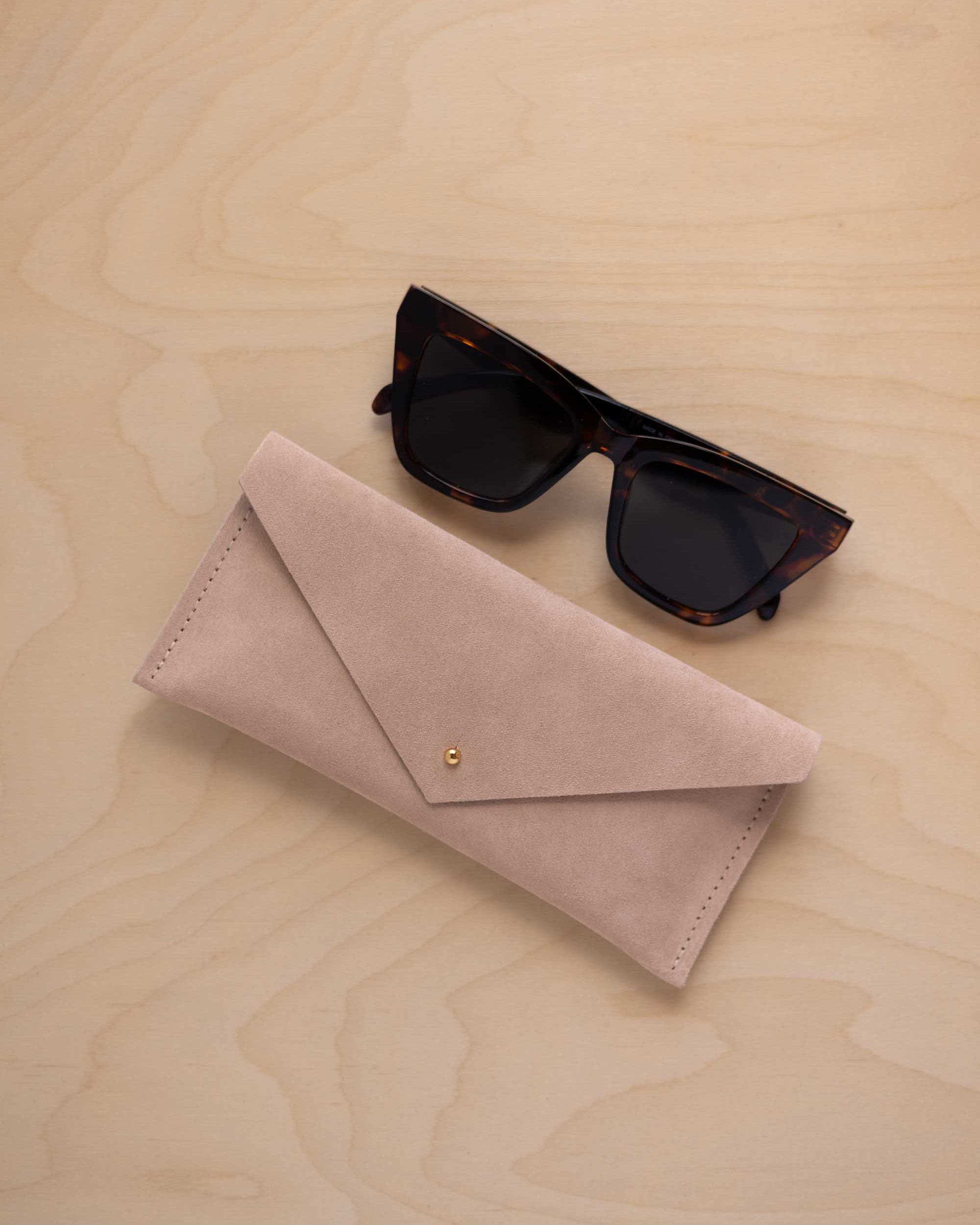 A pair of dark tortoiseshell sunglasses sit next to a warm beige suede envelope style eye glasses case with a small gold button, set on a light wood grain background.