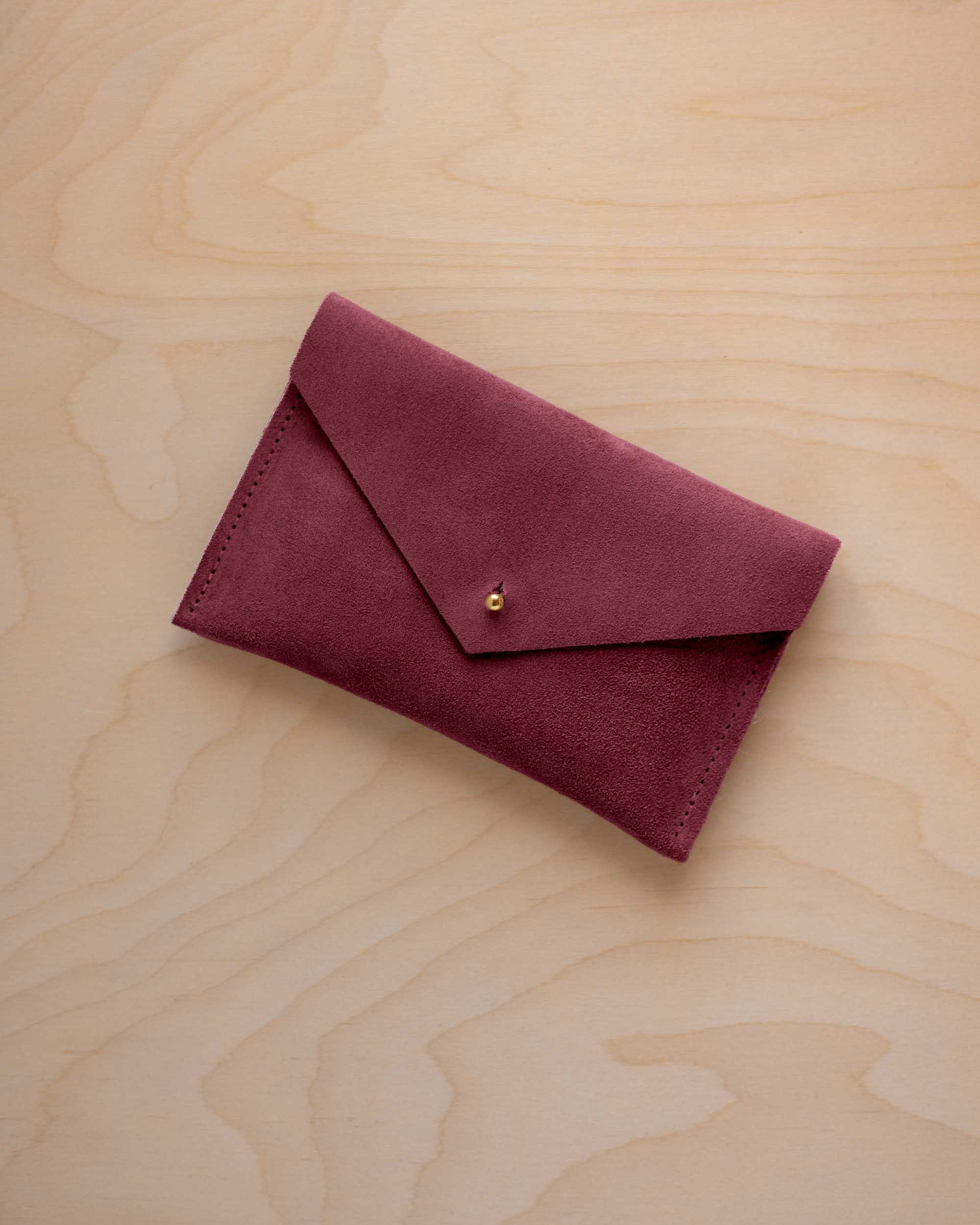 A small burgundy coloured suede envelope-style pouch with a gold button, resting on a light wood grain surface.