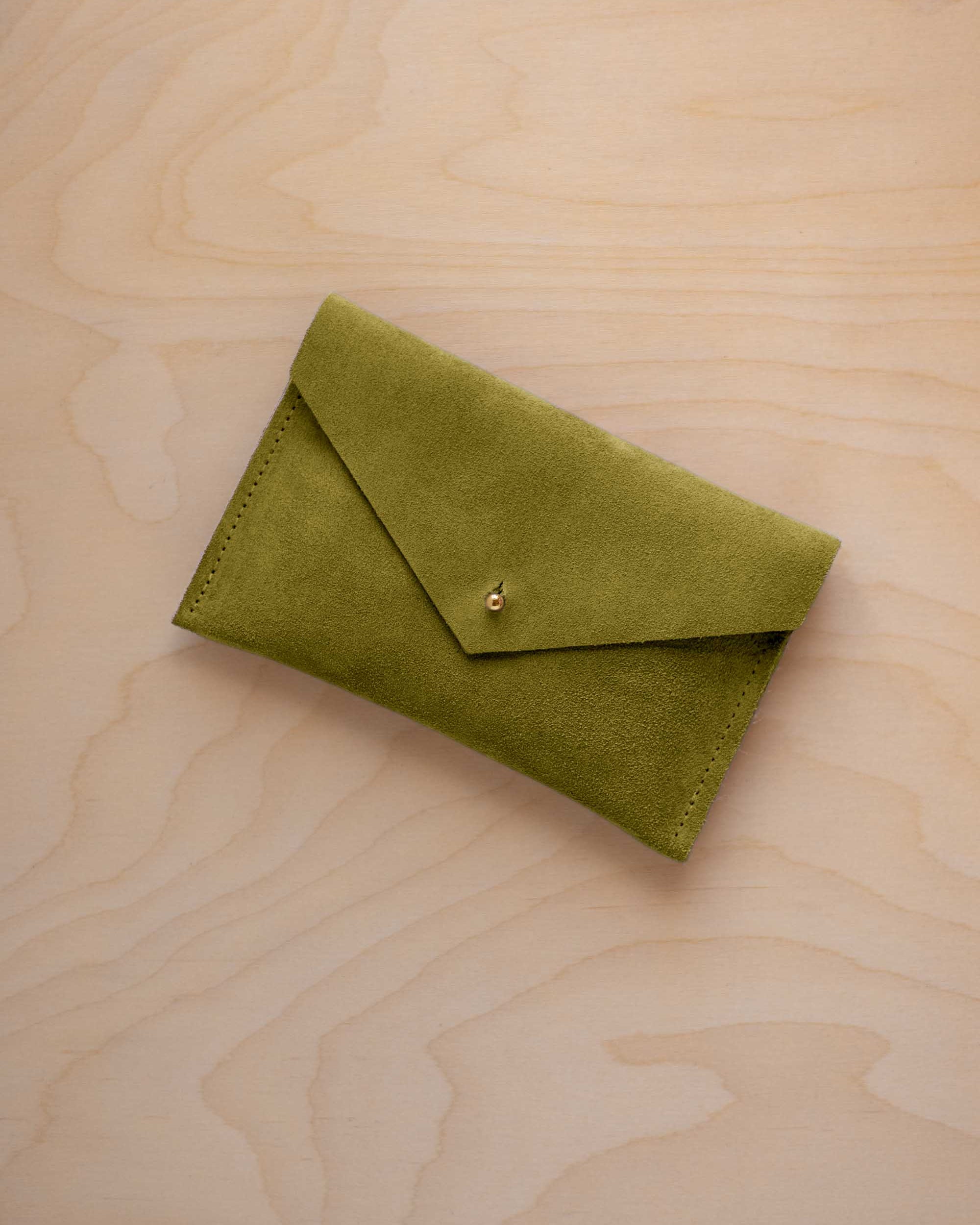 A small chartreuse green coloured suede envelope-style pouch with a gold button, resting on a light wood grain surface.