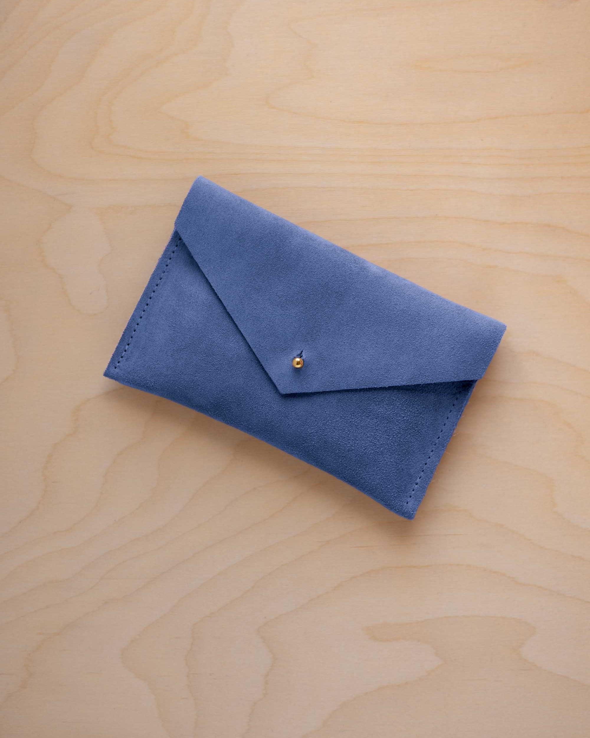 A small cobalt blue coloured suede envelope-style pouch with a gold button, resting on a light wood grain surface.
