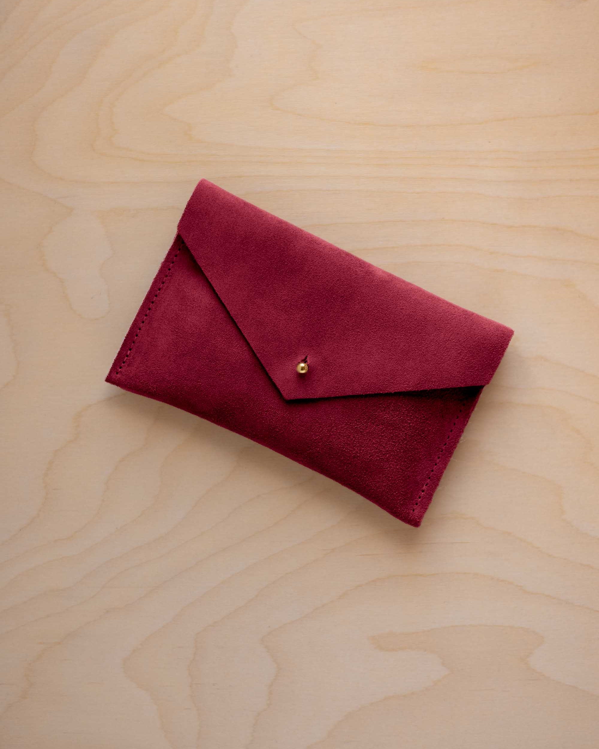 A small crimson red coloured suede envelope-style pouch with a gold button, resting on a light wood grain surface.