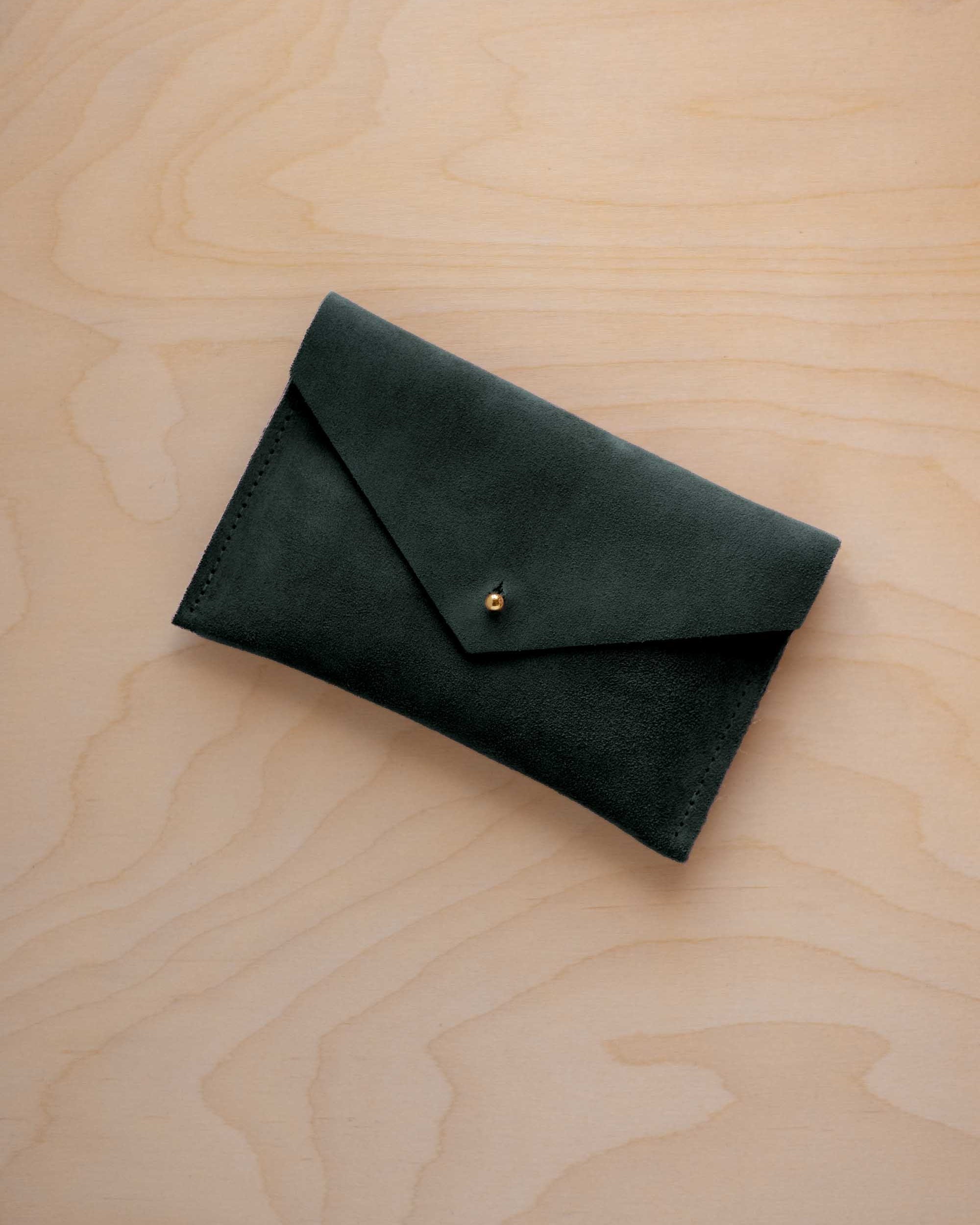 A small deep forest green coloured suede envelope-style pouch with a gold button, resting on a light wood grain surface.