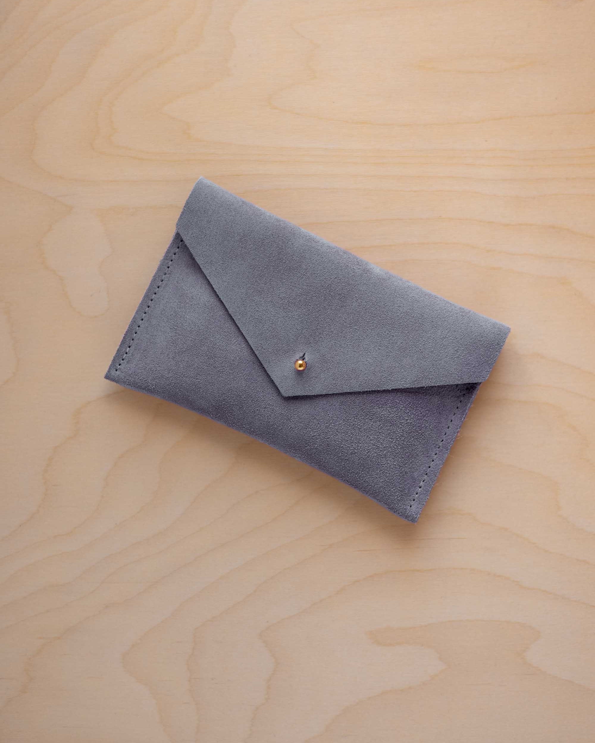A small dusky blue coloured suede envelope-style pouch with a gold button, resting on a light wood grain surface.