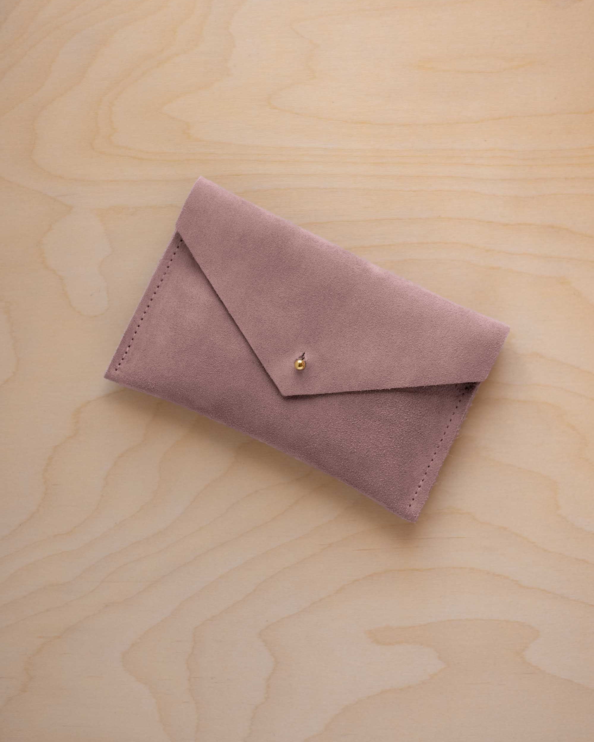 A small dusty mauve coloured suede envelope-style pouch with a gold button, resting on a light wood grain surface.