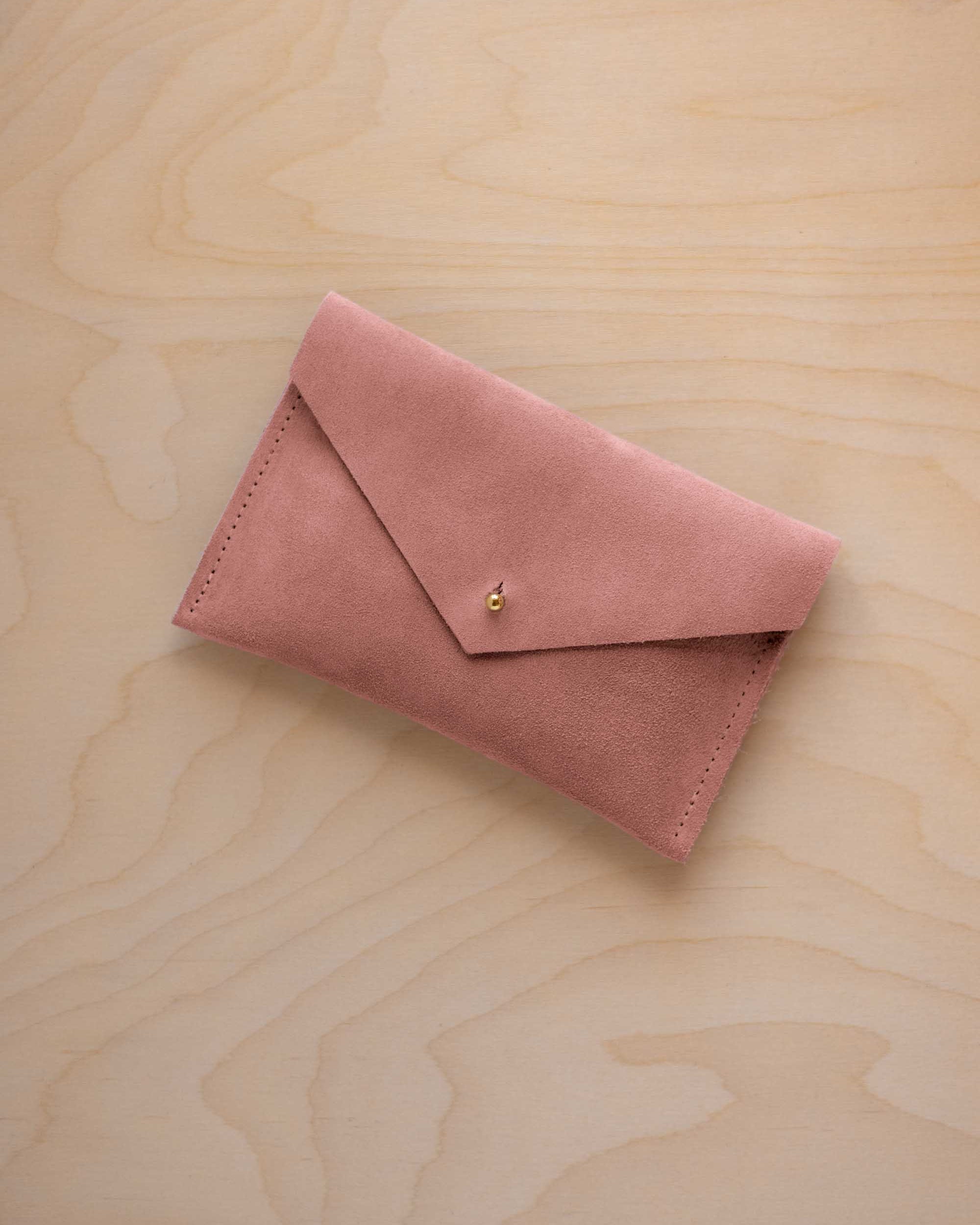A small dusty rose coloured suede envelope-style pouch with a gold button, resting on a light wood grain surface.