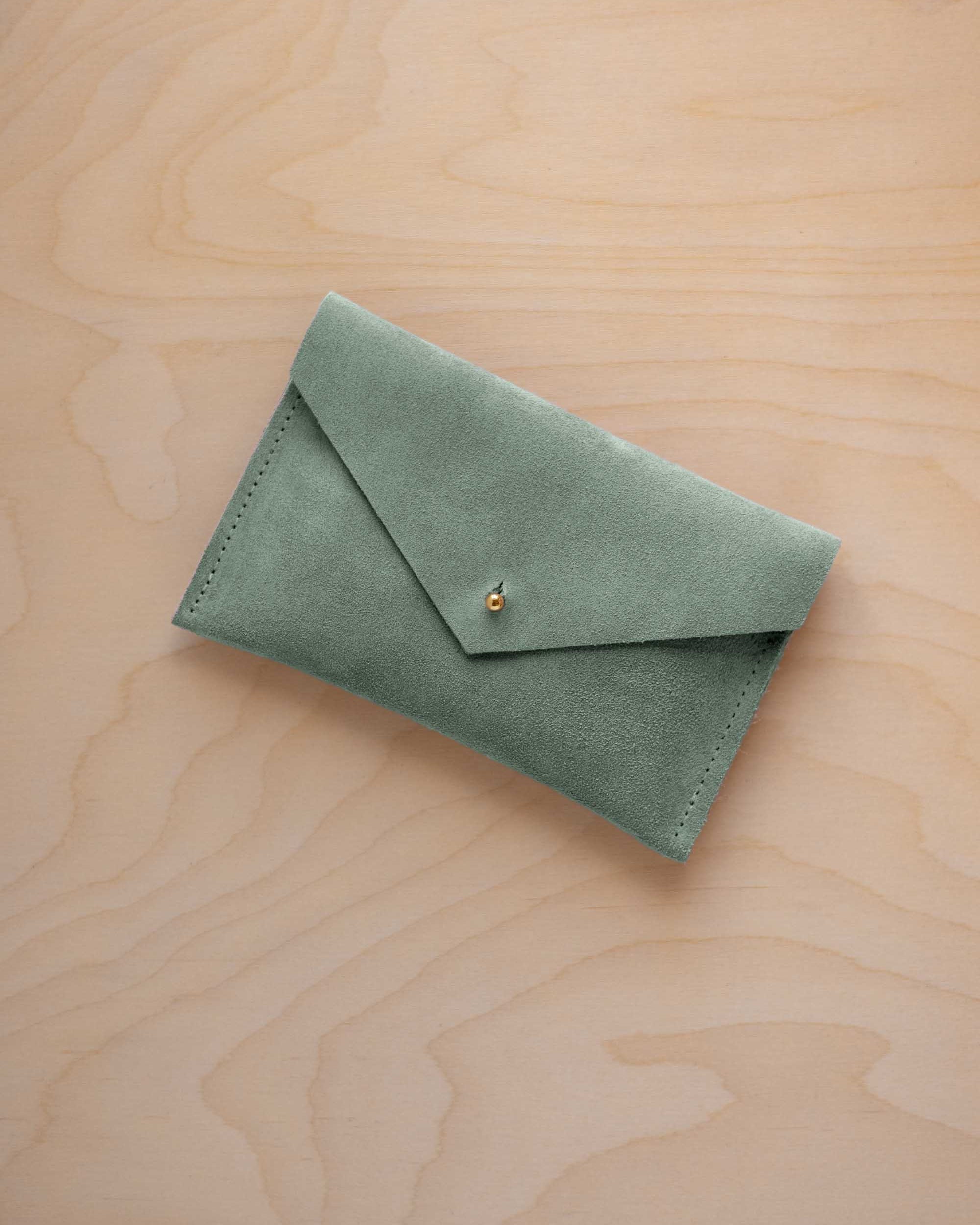 A small eucalyptus green coloured suede envelope-style pouch with a gold button, resting on a light wood grain surface.