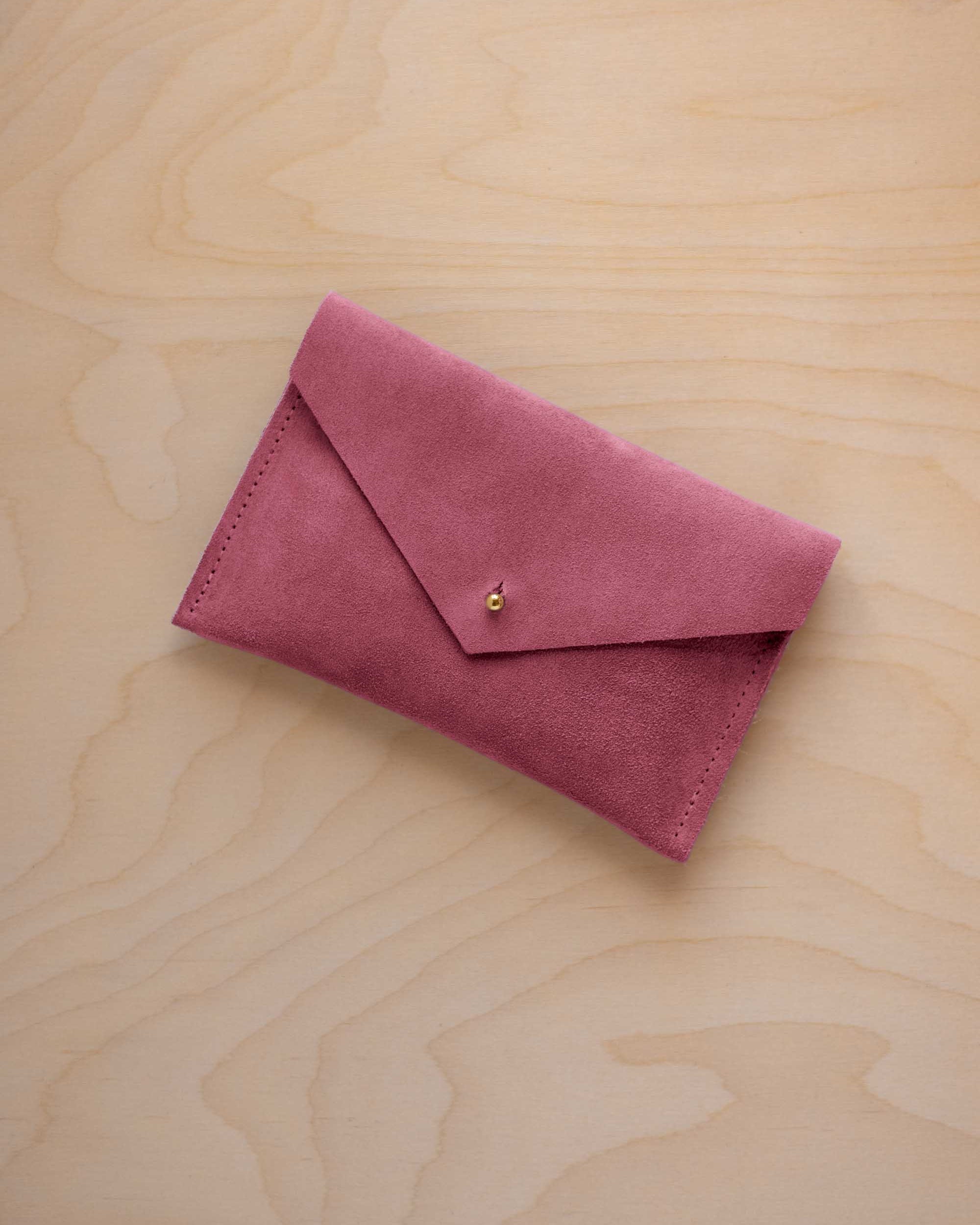 A small faded rose coloured suede envelope-style pouch with a gold button, resting on a light wood grain surface.