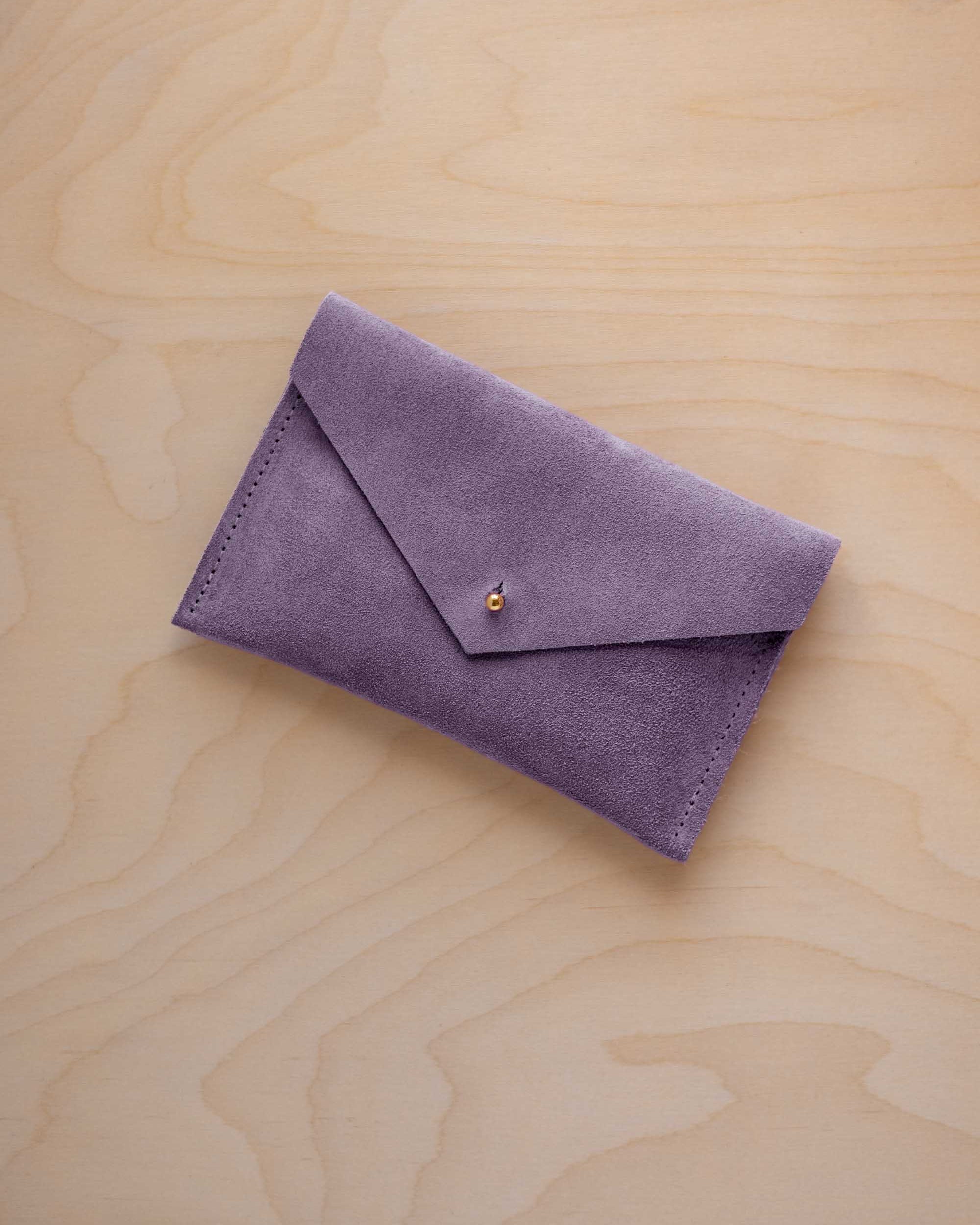 A small lavender coloured suede envelope-style pouch with a gold button, resting on a light wood grain surface.