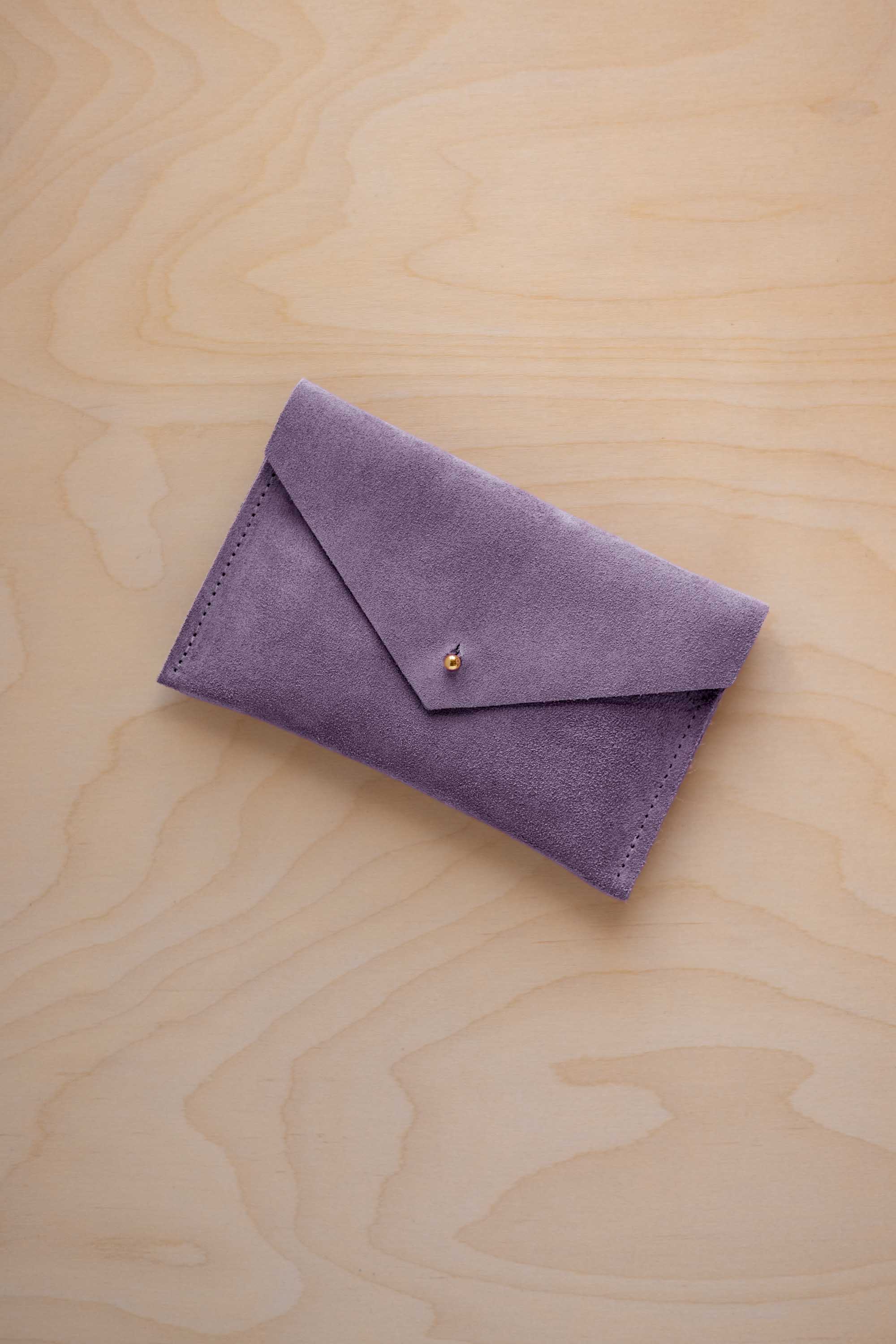 A small lavender coloured suede envelope-style pouch with a gold button, resting on a light wood grain surface.