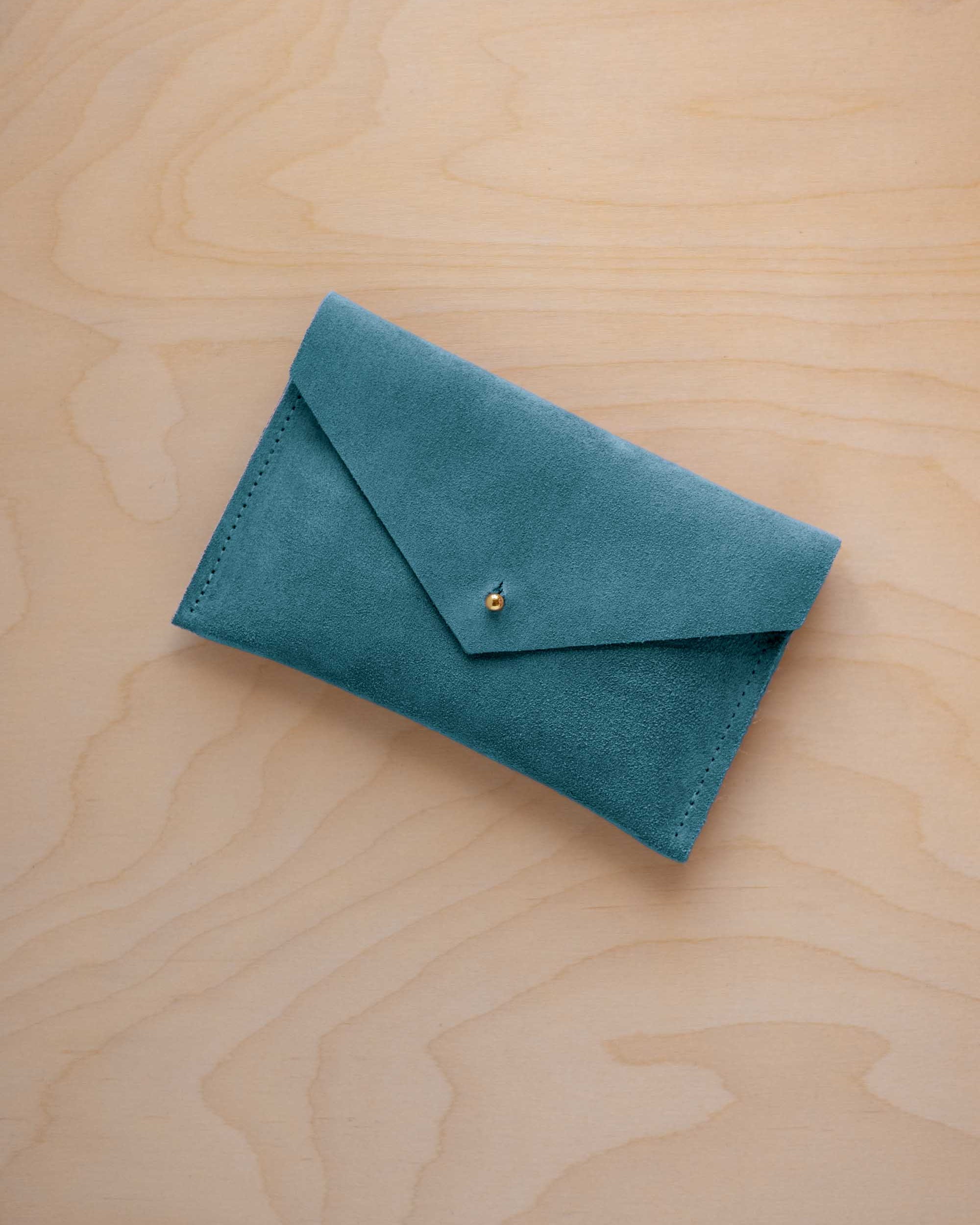 A small marine teal coloured suede envelope-style pouch with a gold button, resting on a light wood grain surface.
