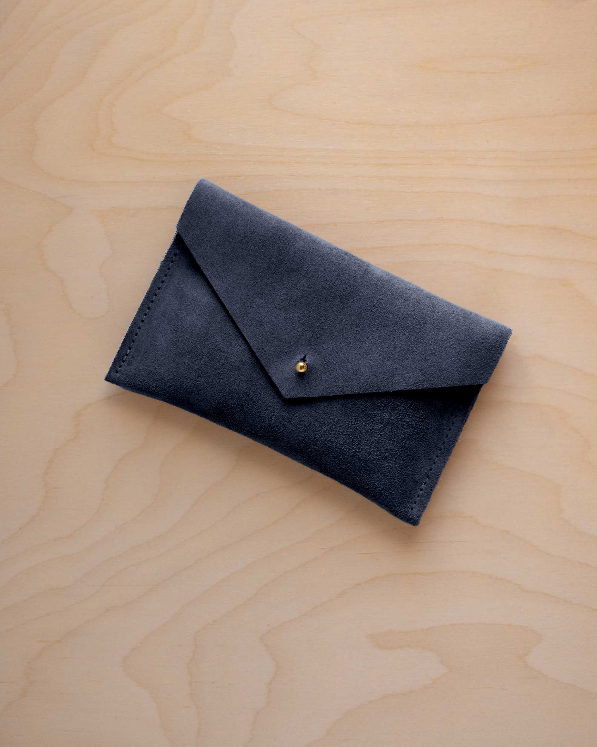 A small midnight navy blue coloured suede envelope-style wallet pouch with a gold button, resting on a light wood grain surface.