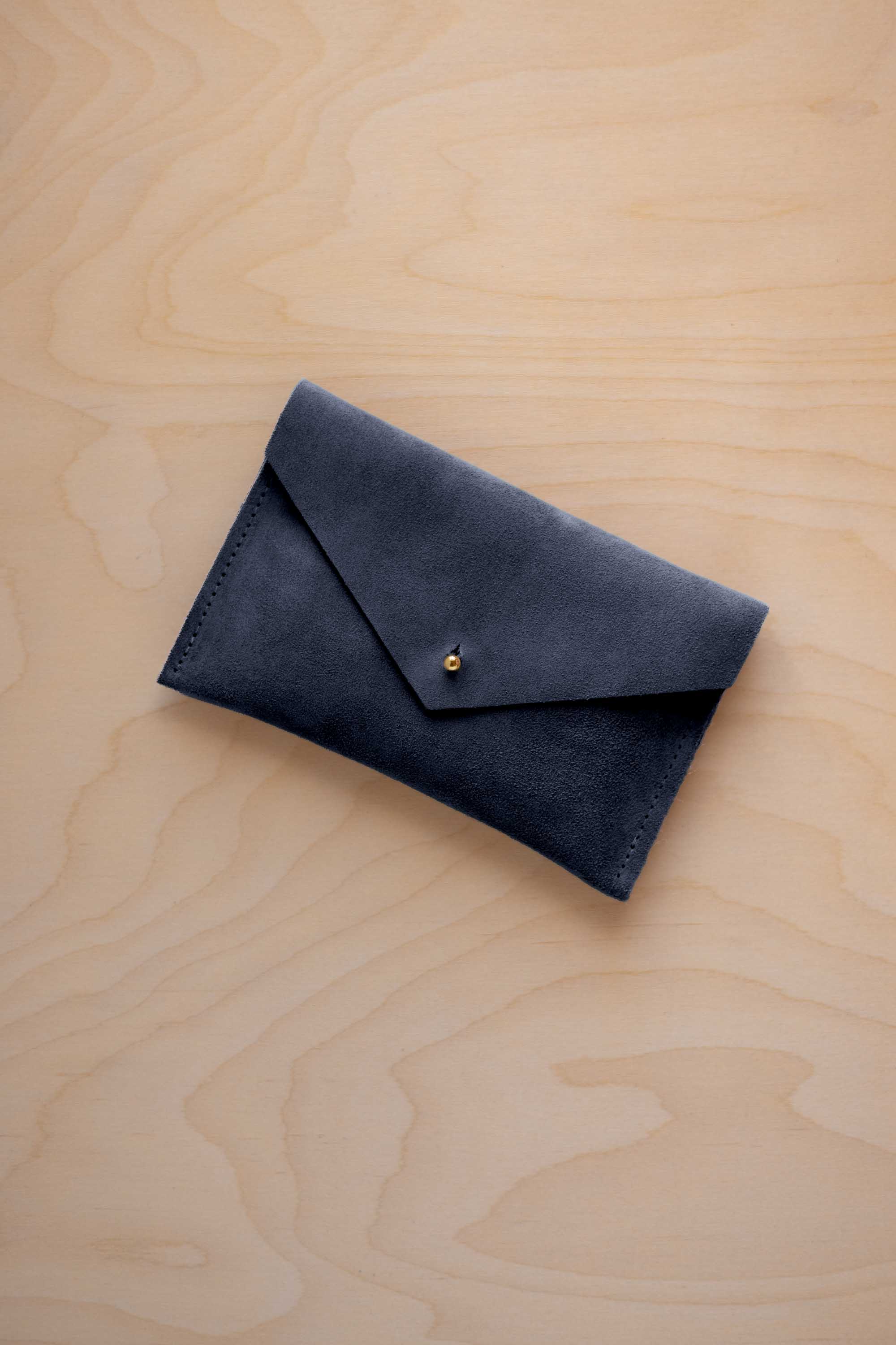 A small midnight navy blue coloured suede envelope-style wallet pouch with a gold button, resting on a light wood grain surface.