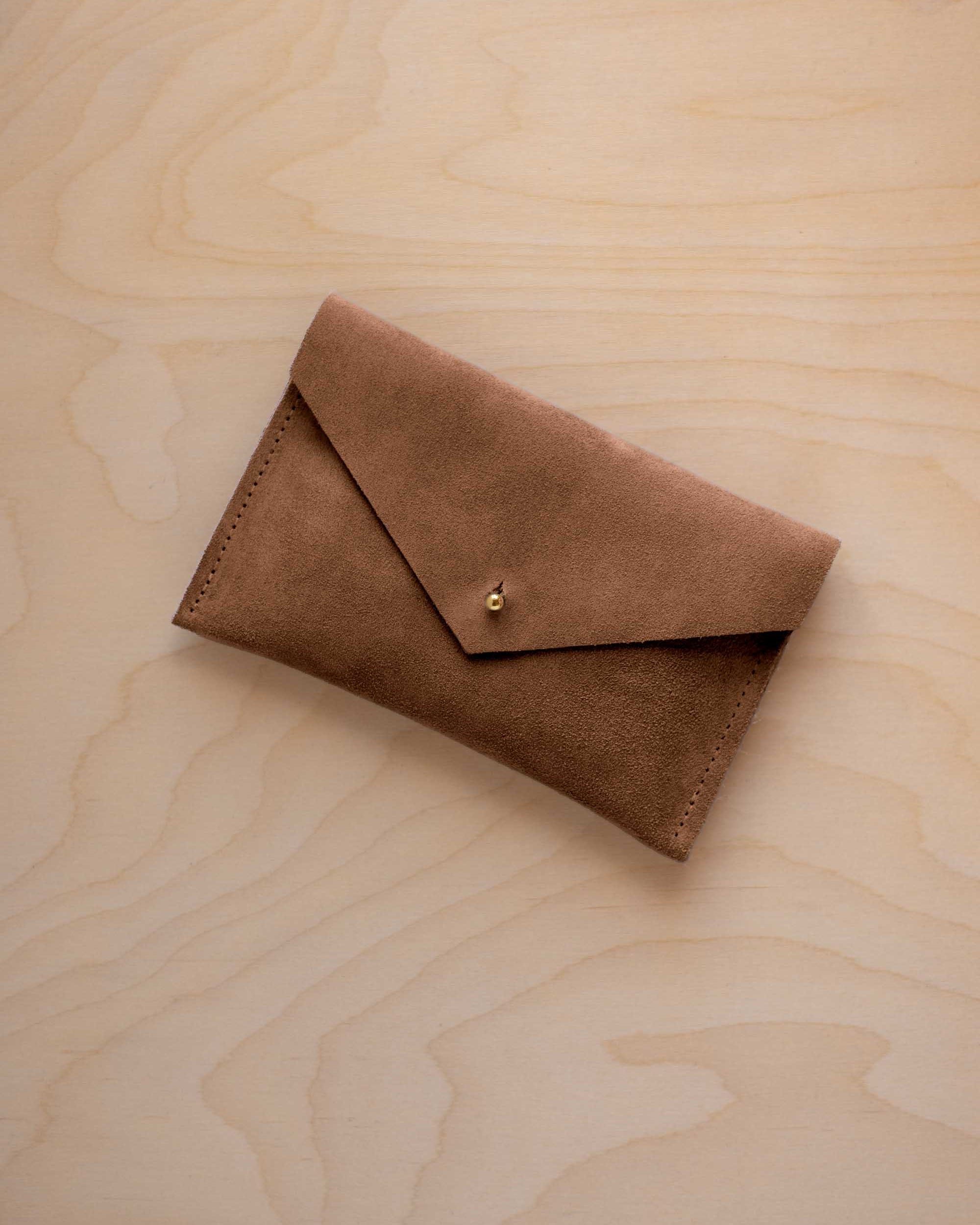 A small mocha brown coloured suede envelope-style pouch with a gold button, resting on a light wood grain surface.