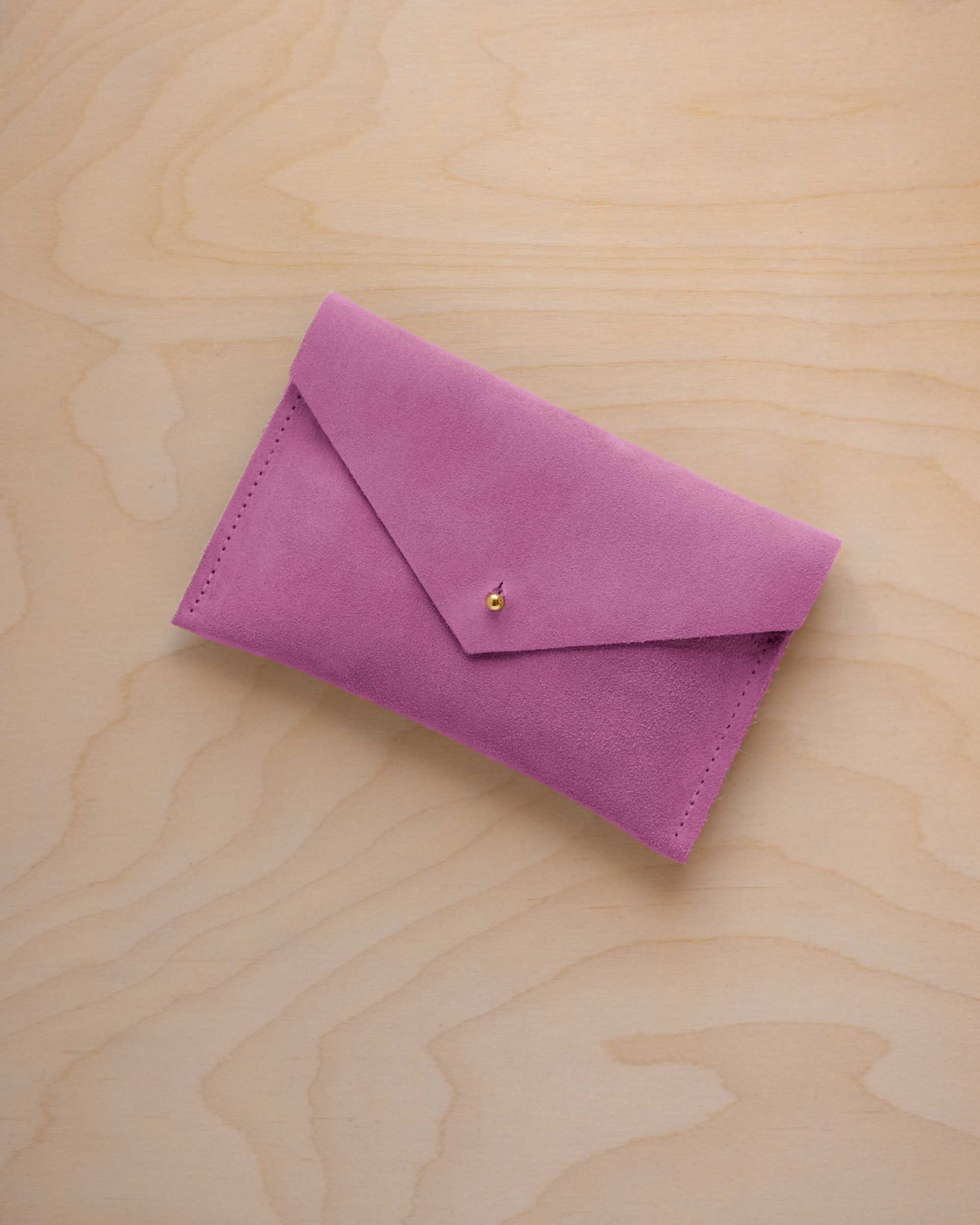 A small peony pink coloured suede envelope-style pouch with a gold button, resting on a light wood grain surface.