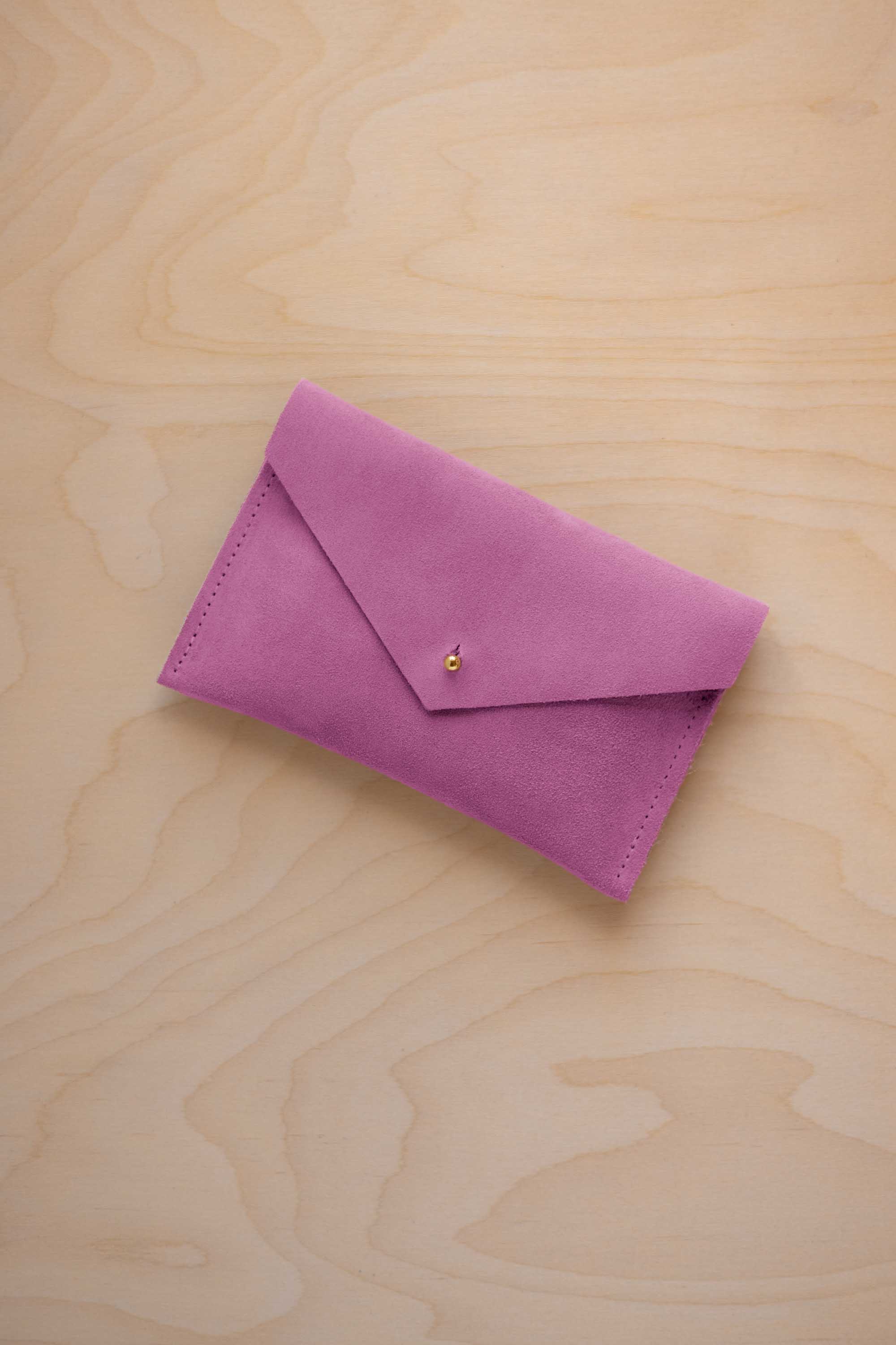 A small peony pink coloured suede envelope-style pouch with a gold button, resting on a light wood grain surface.