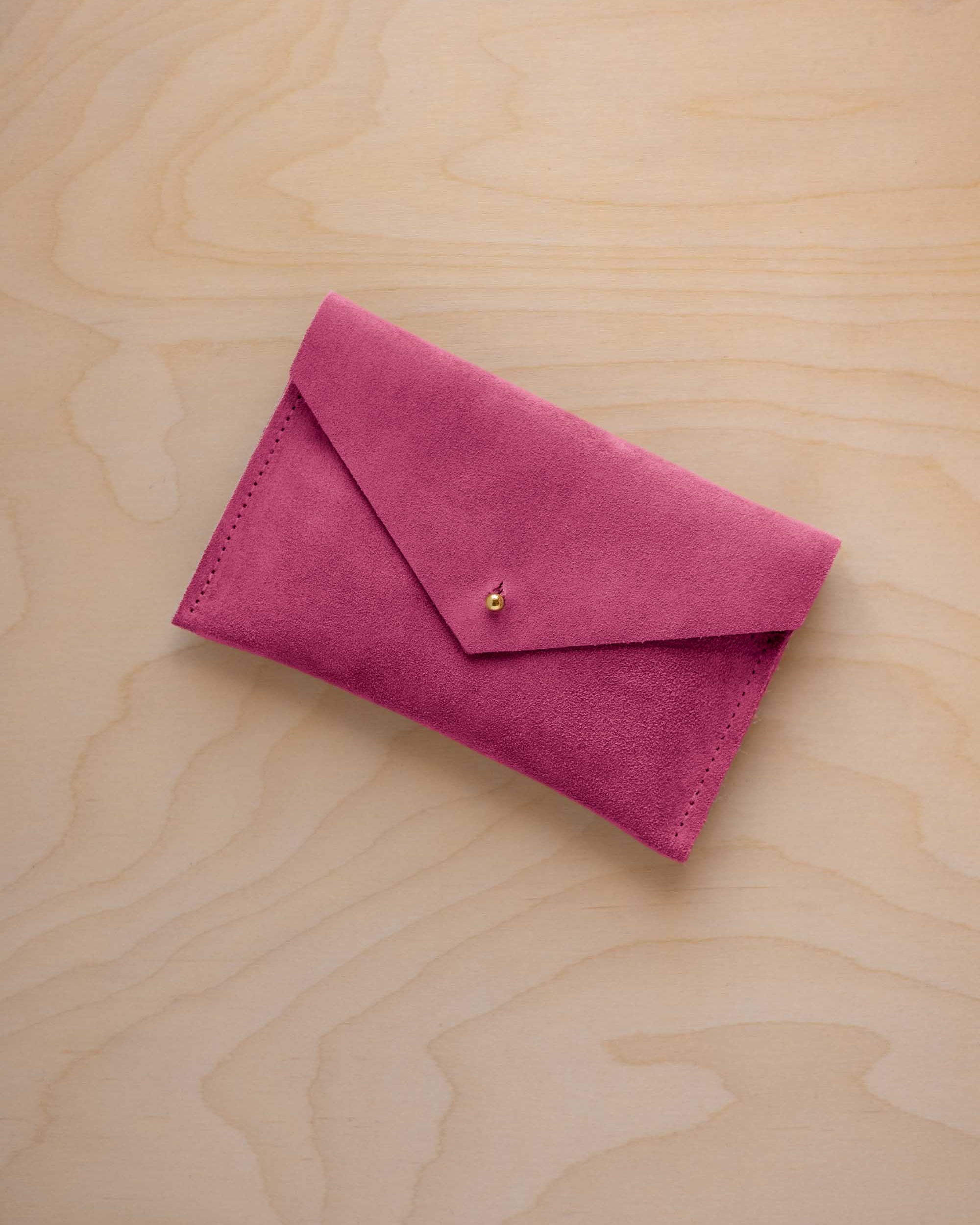 A small raspberry coloured suede envelope-style pouch with a gold button, resting on a light wood grain surface.