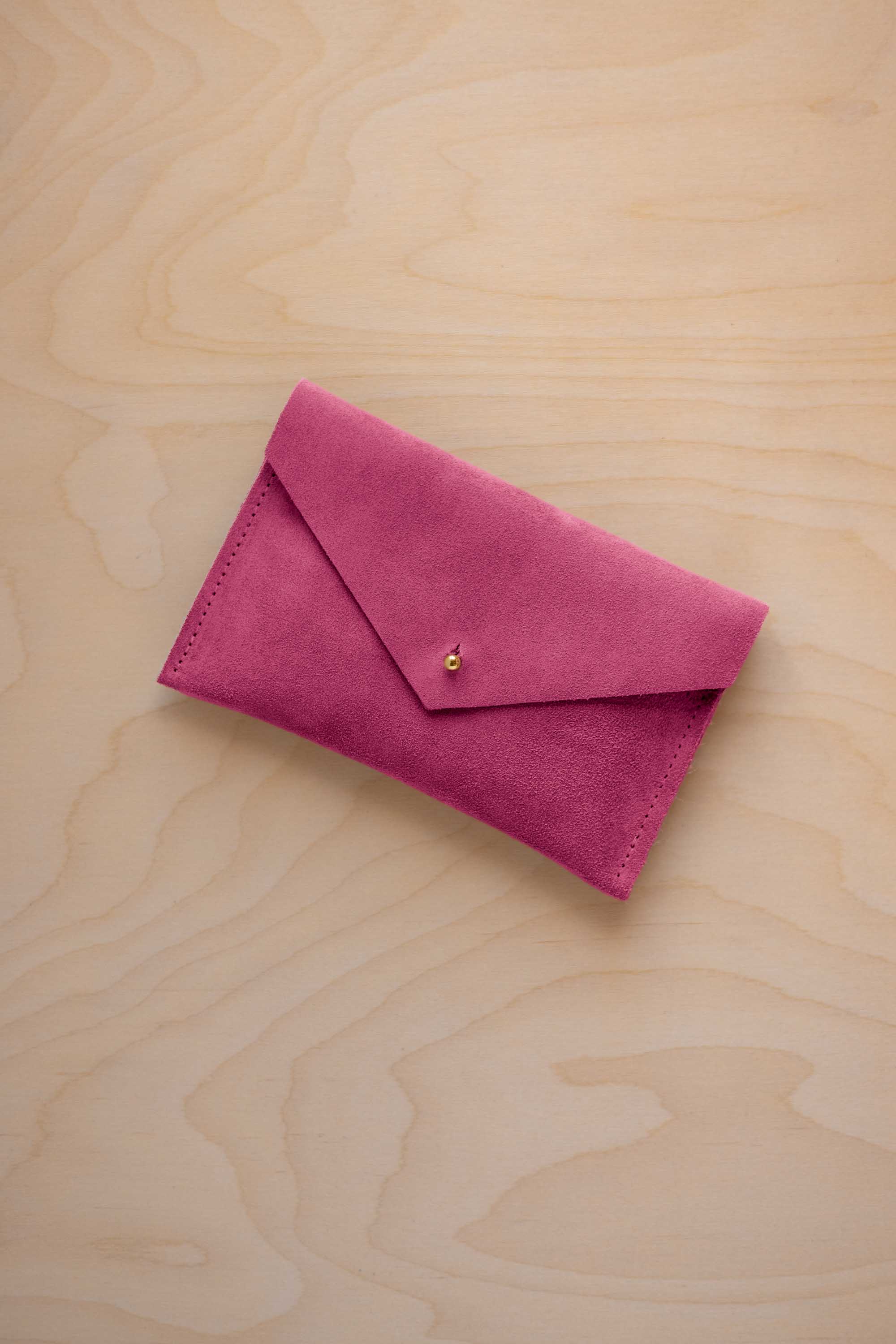 A small raspberry coloured suede envelope-style pouch with a gold button, resting on a light wood grain surface.