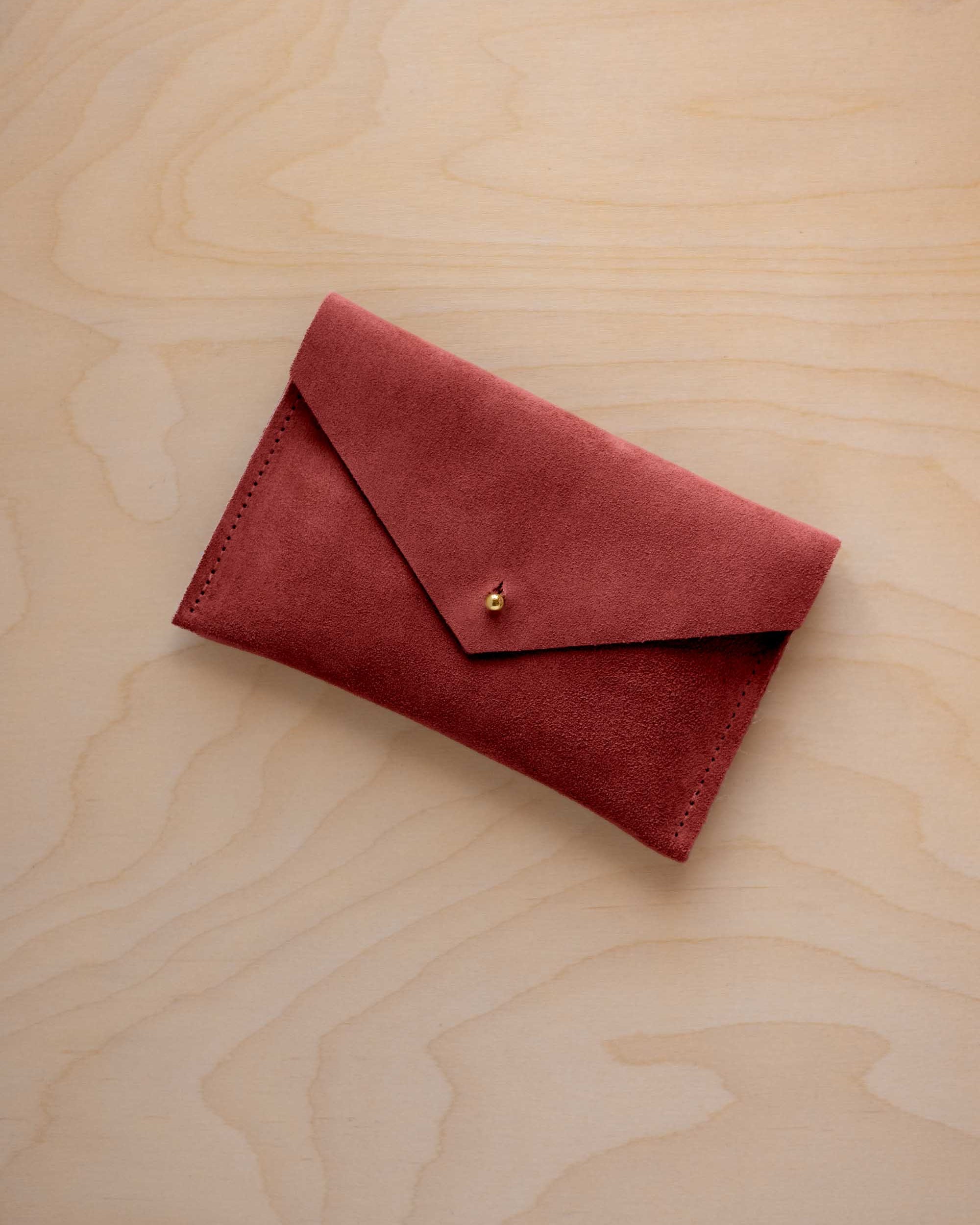 A small rust red coloured suede envelope-style wallet pouch with a gold button, resting on a light wood grain surface.
