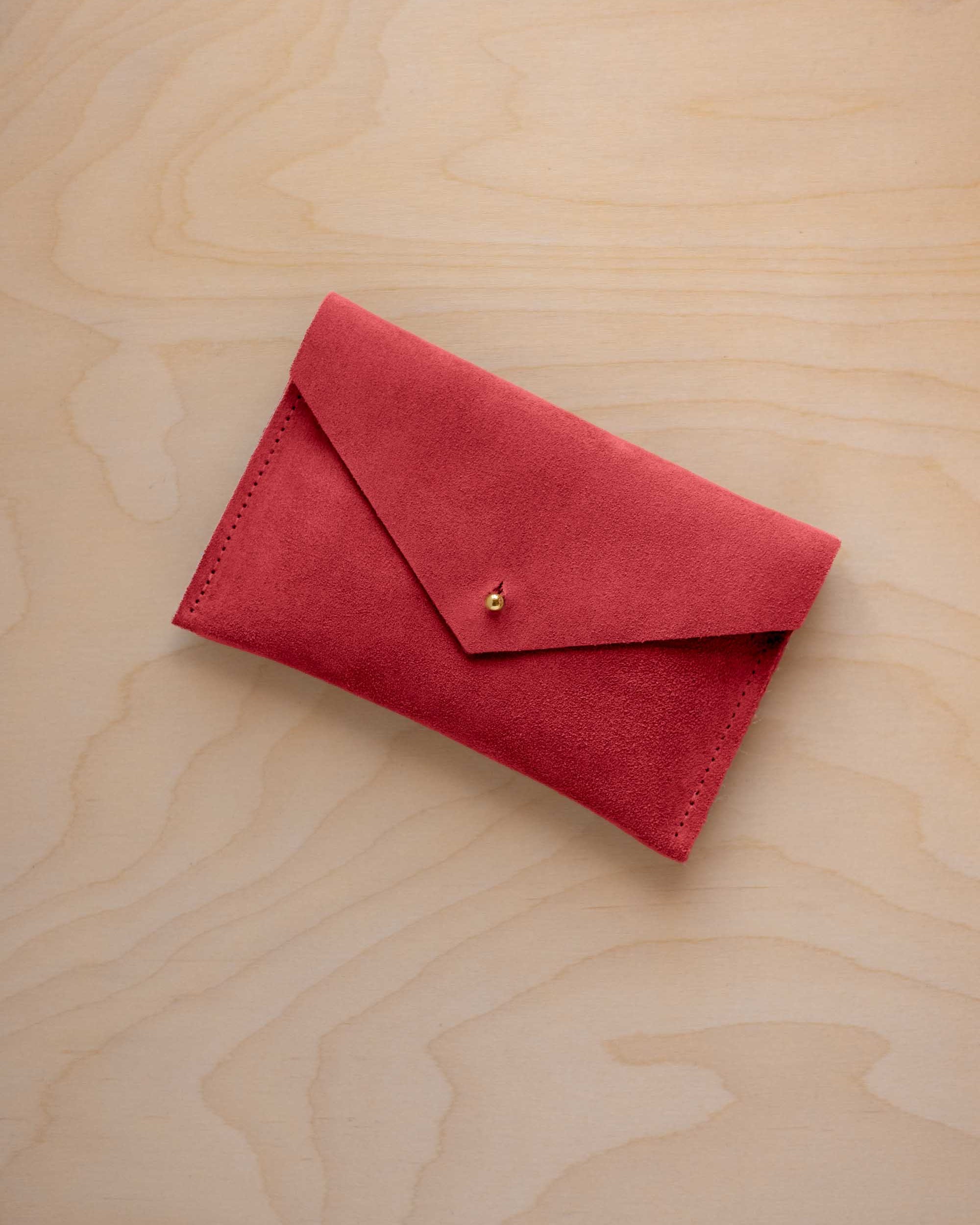 A small scarlet red coloured suede envelope-style pouch with a gold button, resting on a light wood grain surface.
